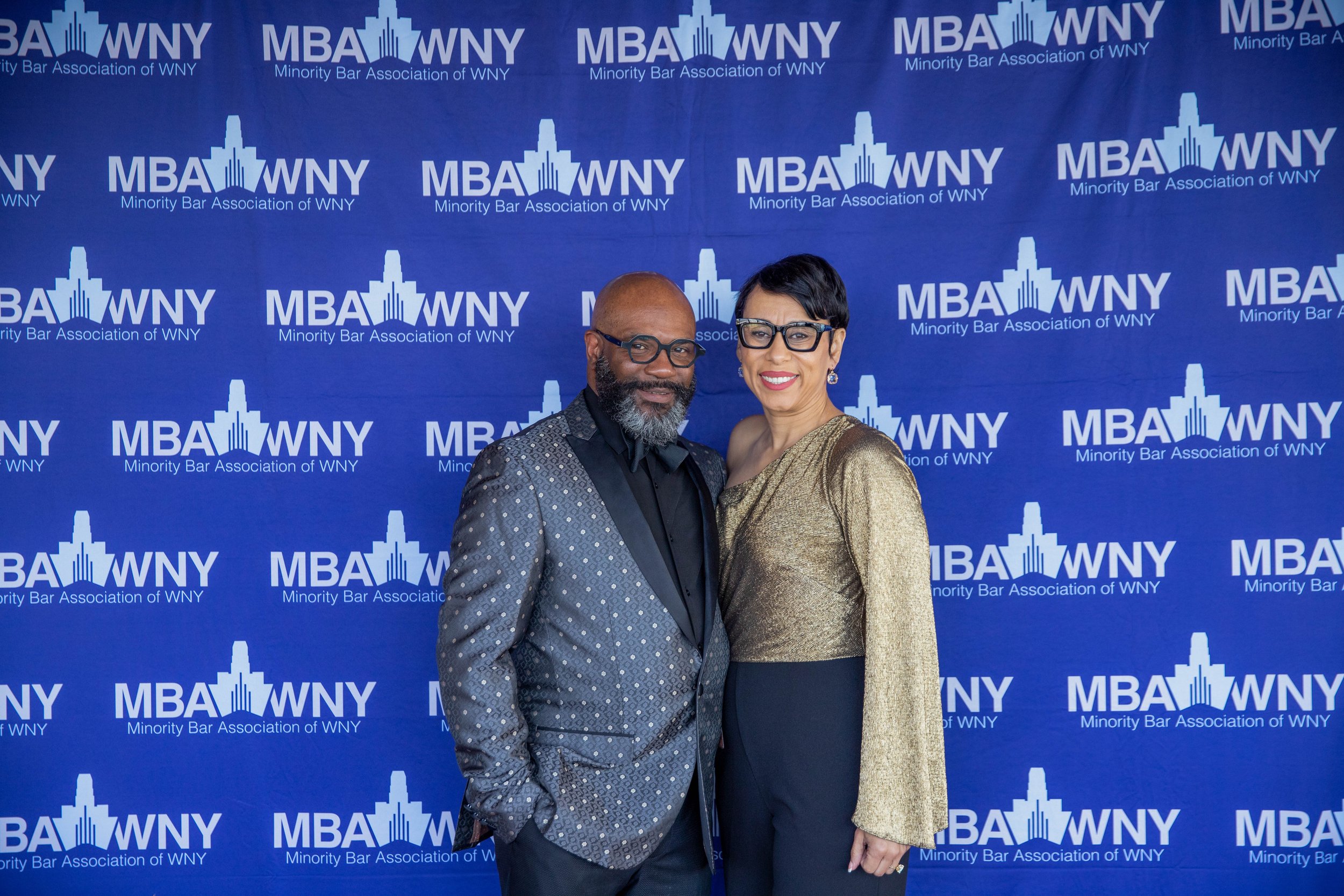 Two people dressed in formal attire standing in front of a blue backdrop with the logo and text of the Minority Bar Association of WNY, smiling at the camera.