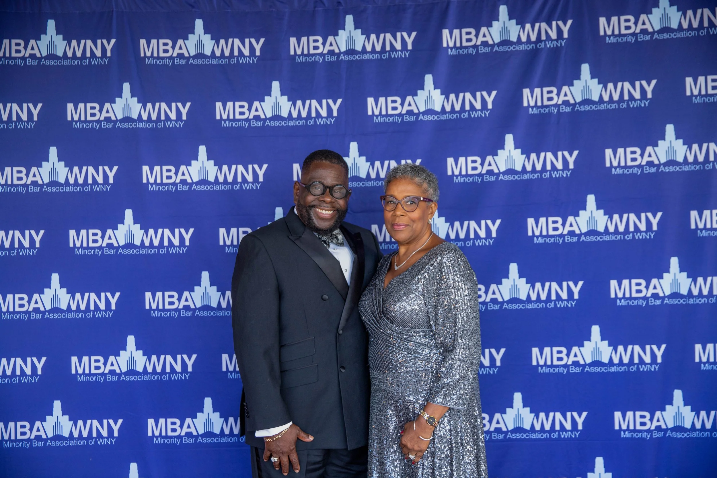 Two individuals, a man and a woman, standing in front of a blue backdrop with the logo and text for the Minority Bar Association of WNY, smiling at the camera.