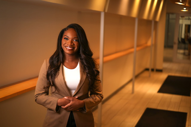 A smiling woman with long dark hair wearing a tan blazer and white top standing in a corridor with wooden flooring and modern lighting.