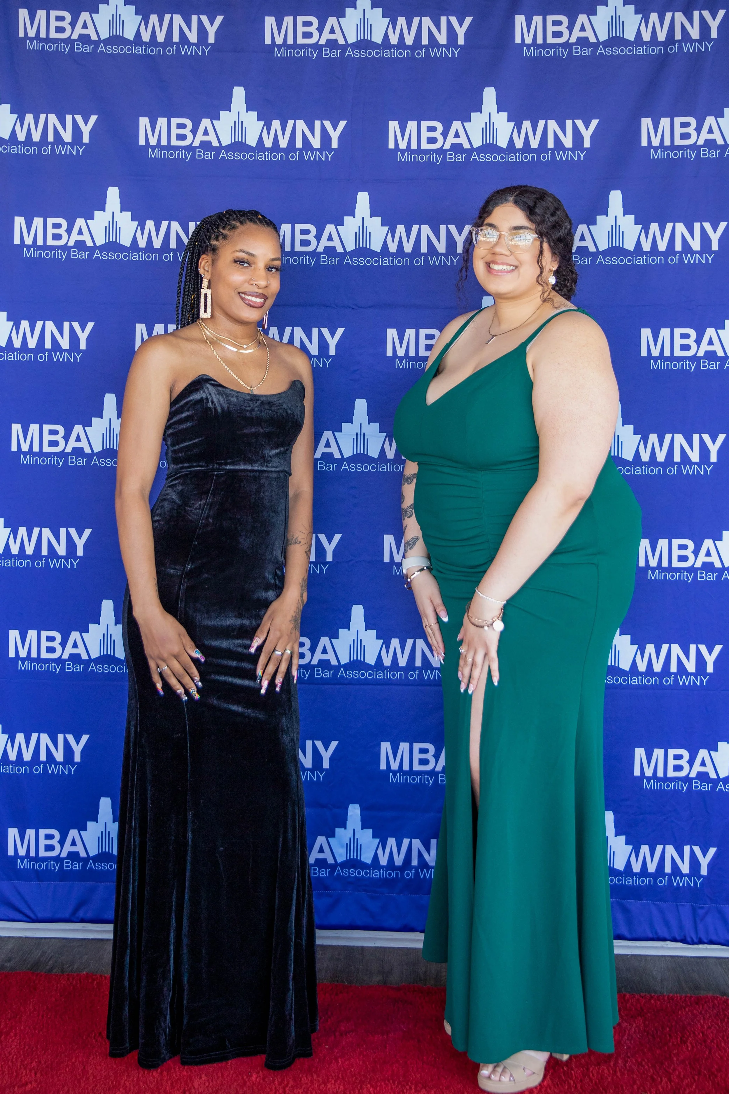 Two women in formal attire standing on a red carpet in front of a blue backdrop with white text and logos for the Minority Bar Association of WNY. One woman is wearing a strapless black velvet dress and the other a green sleeveless gown with a slit. 