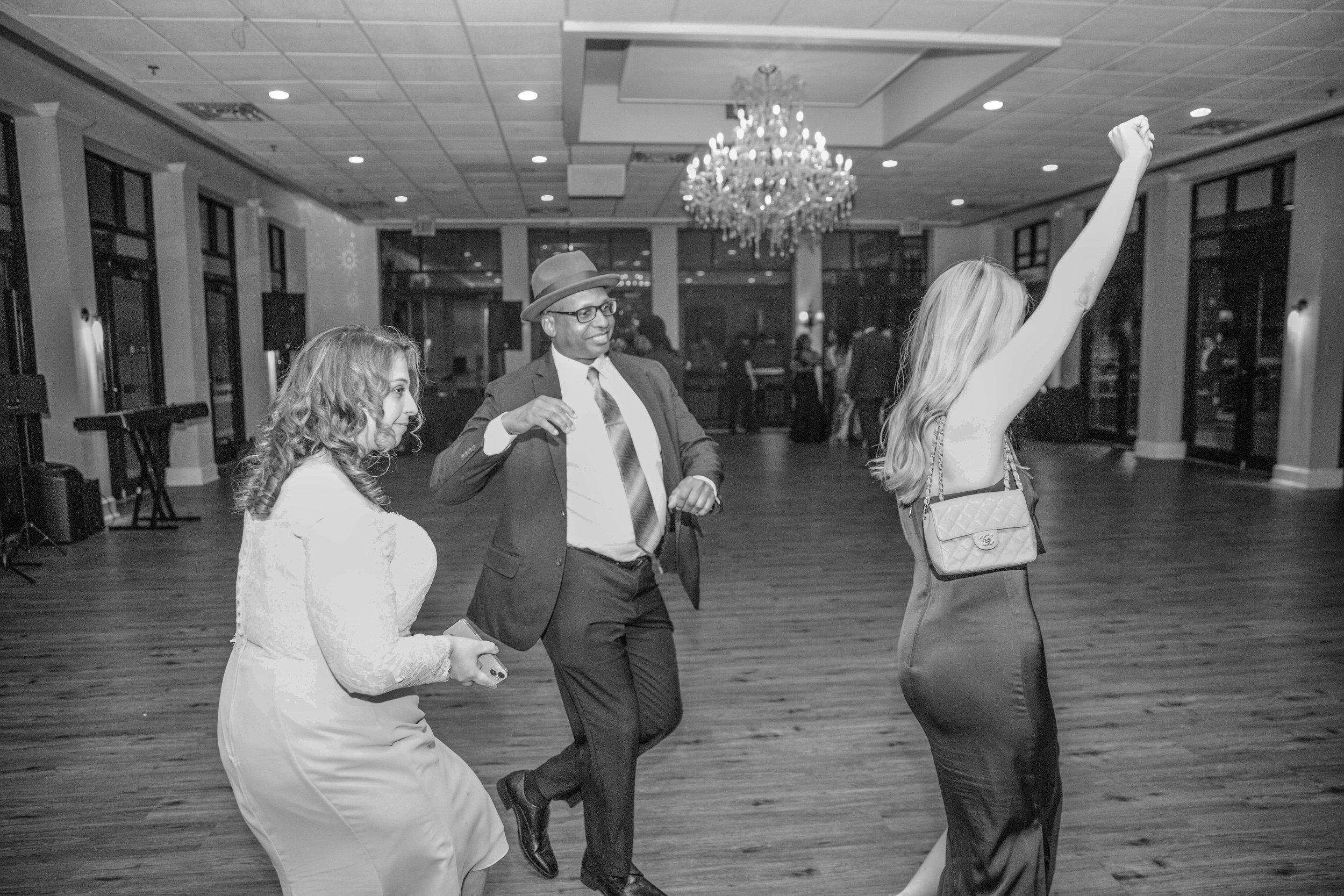People dancing at an indoor event with chandeliers and large windows.