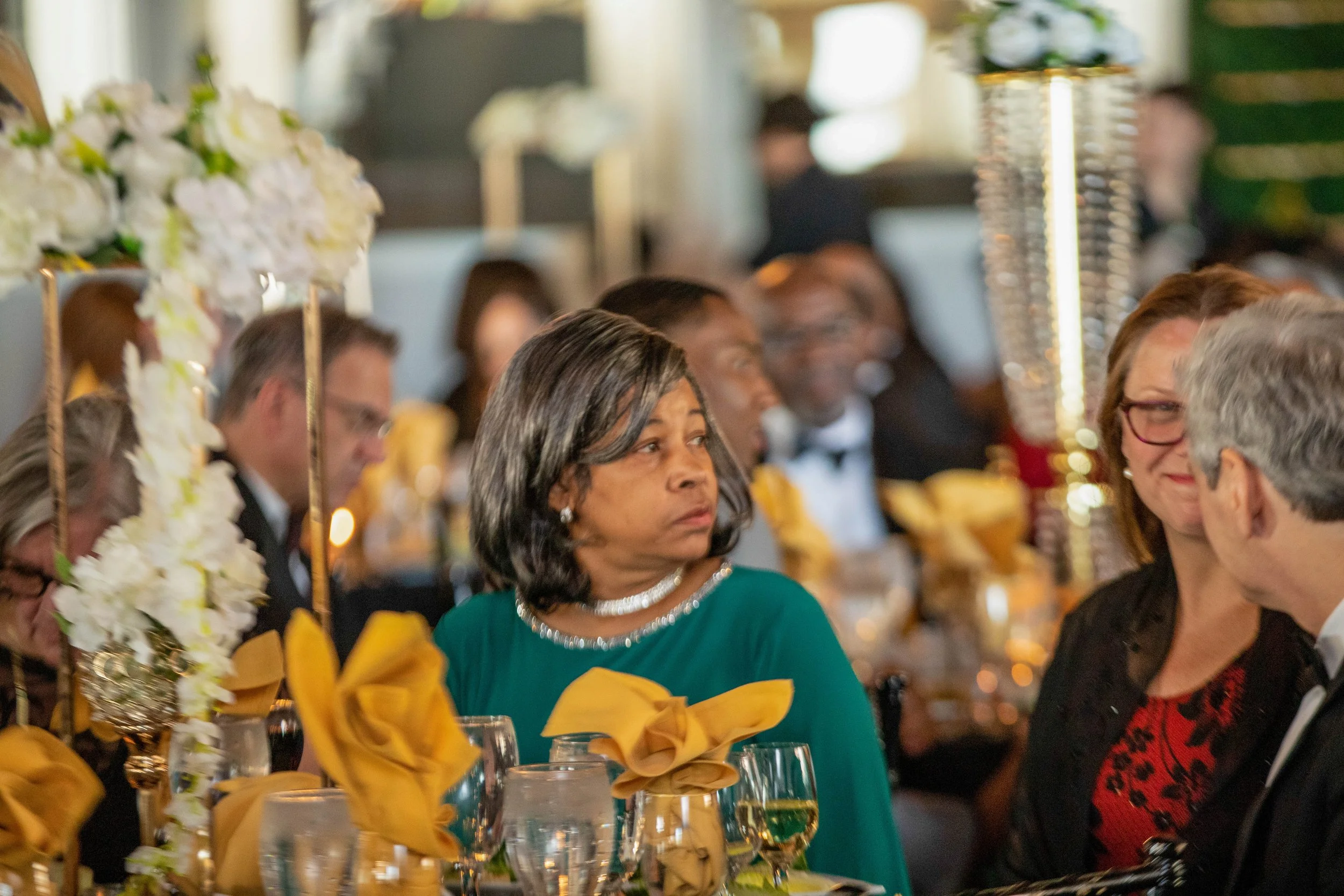 People attending a formal event or banquet, seated at tables with yellow napkins and floral centerpieces.