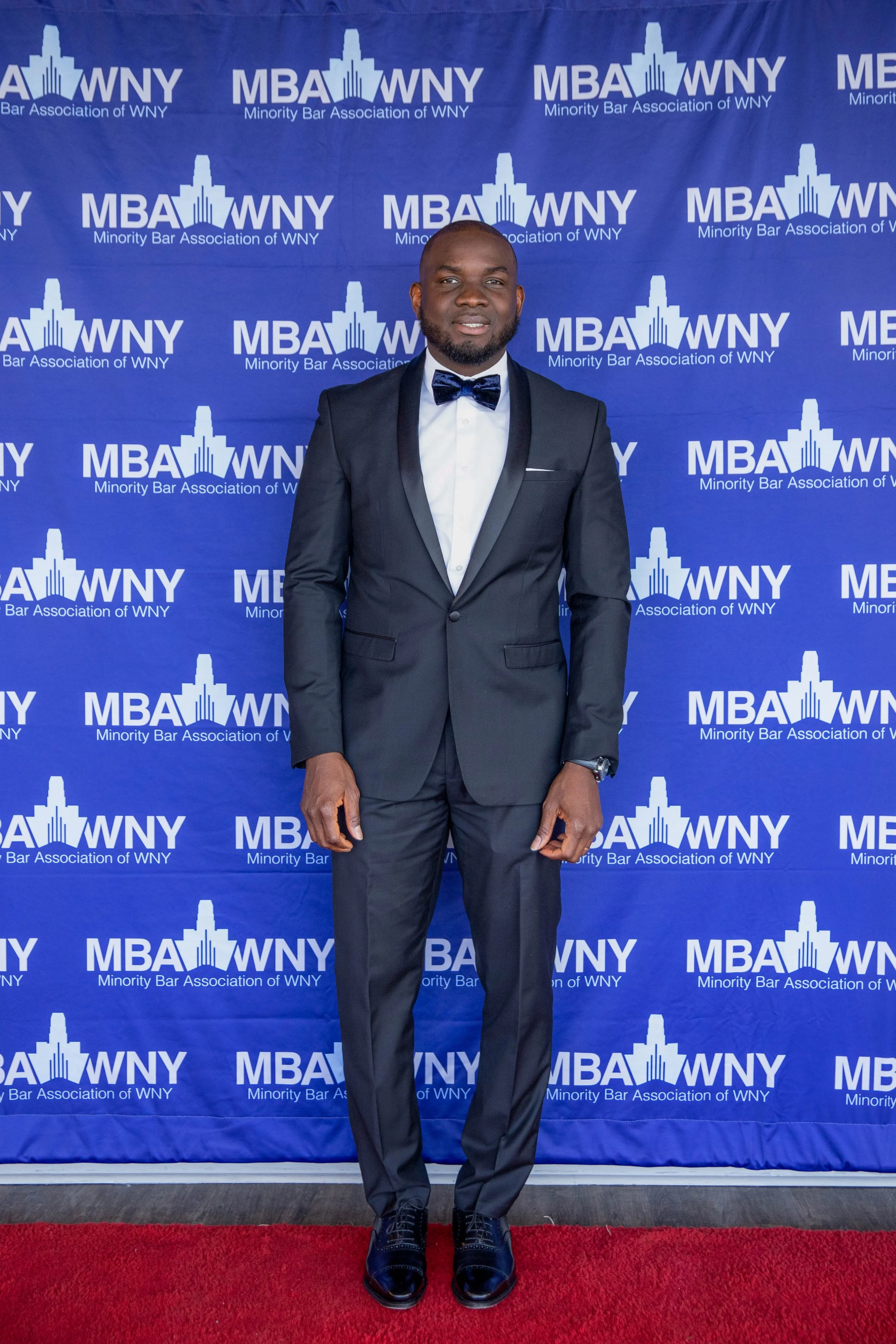 A man wearing a black tuxedo with a white shirt and a black bow tie, standing on a red carpet in front of a backdrop with the text "MBA WNY" and "Minority Bar Association of WNY."