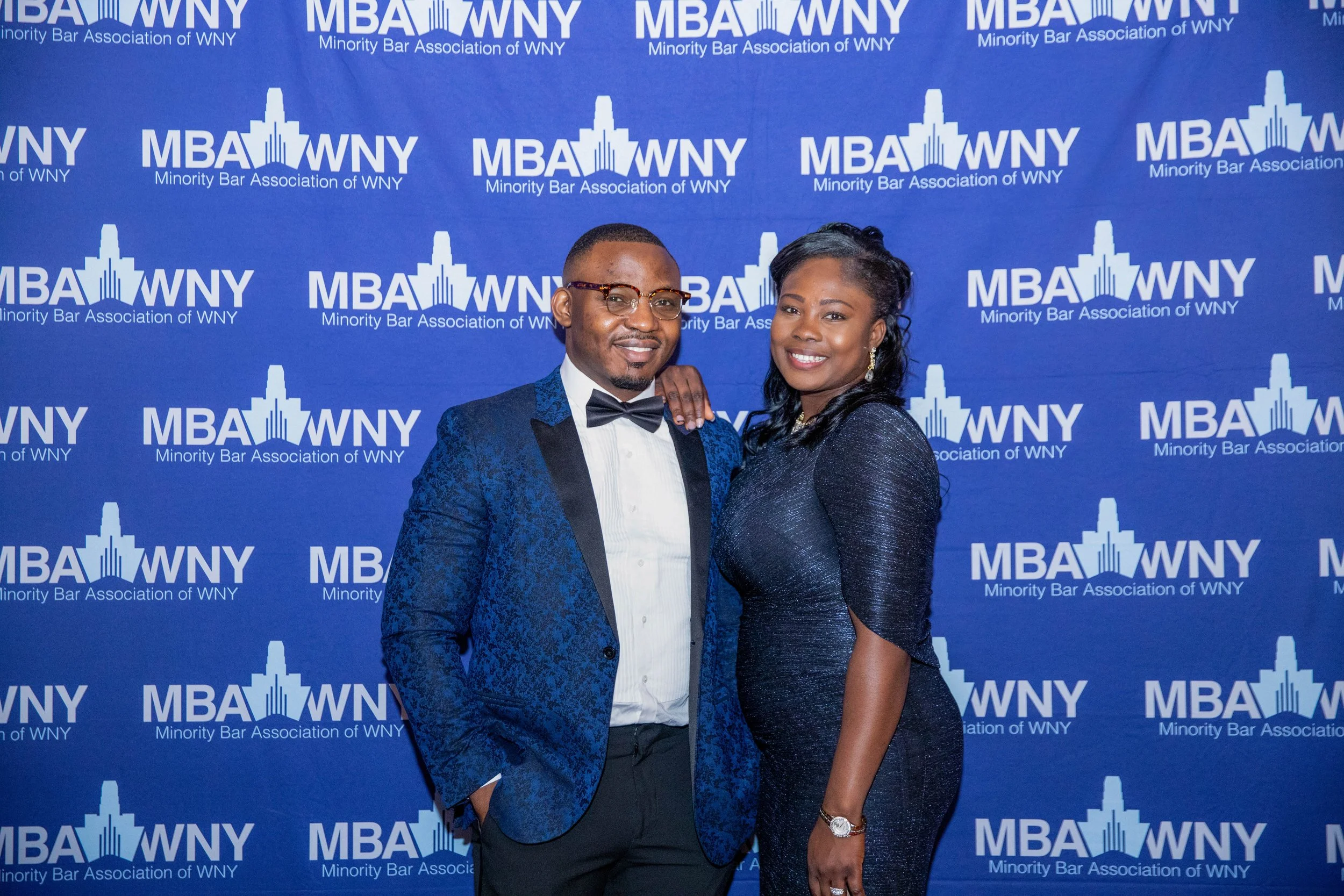A man and woman dressed in formal attire posing in front of a blue backdrop with the logos of the Minority Bar Association of WNY. The man is wearing glasses, a tuxedo, and a bowtie, while the woman is wearing a dark, sparkly dress and earrings.