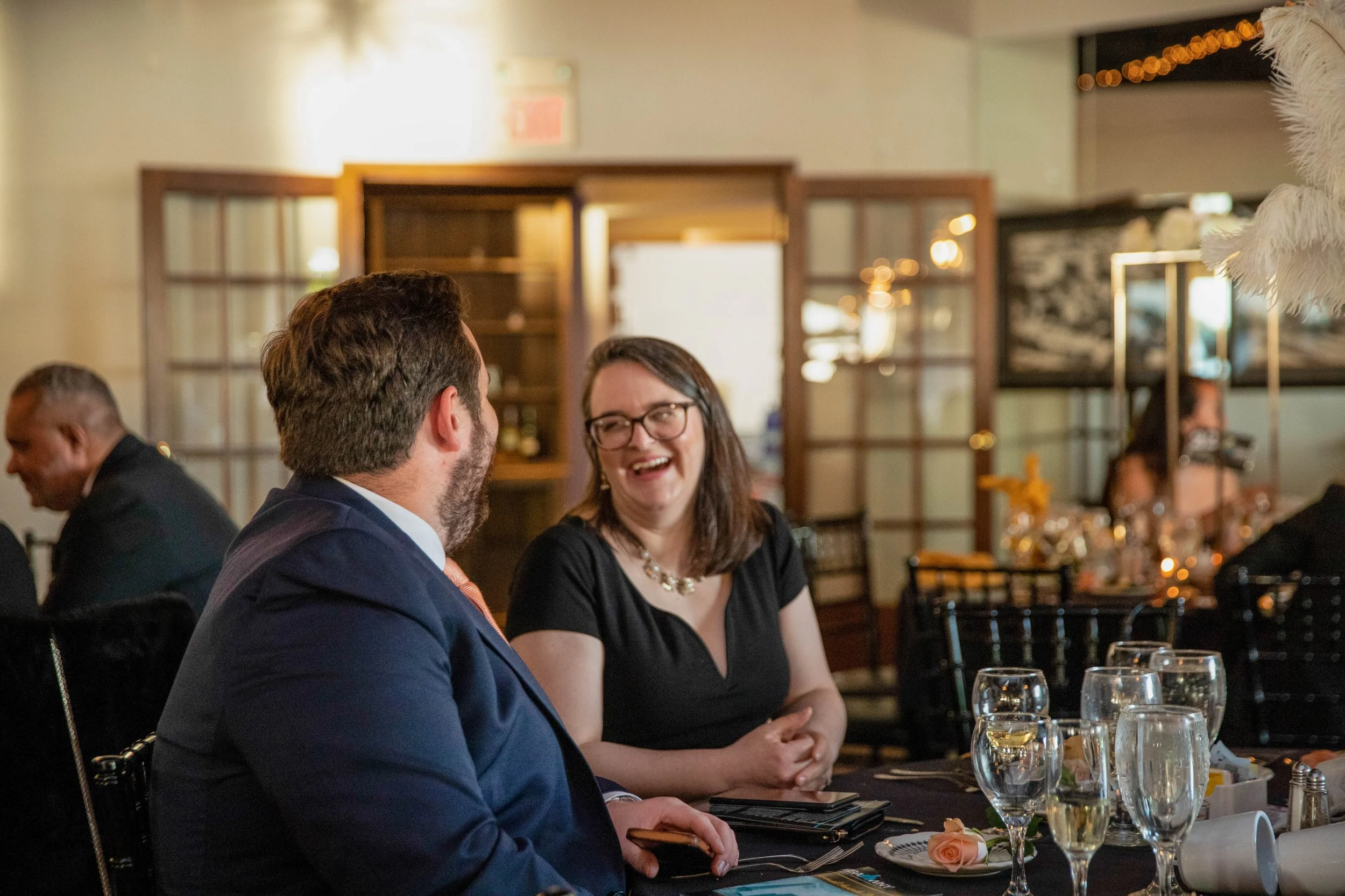 A man and a woman are engaged in conversation at a restaurant table, with several glasses of wine and plates set on the table, and other diners in the background.