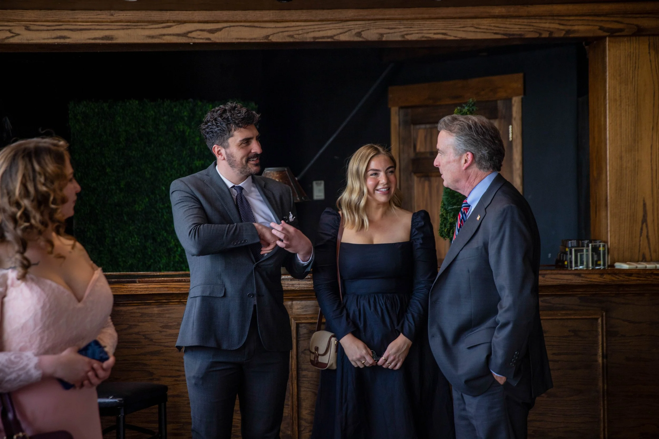 Four people engaged in conversation at a social gathering, with wood-paneled walls in the background.