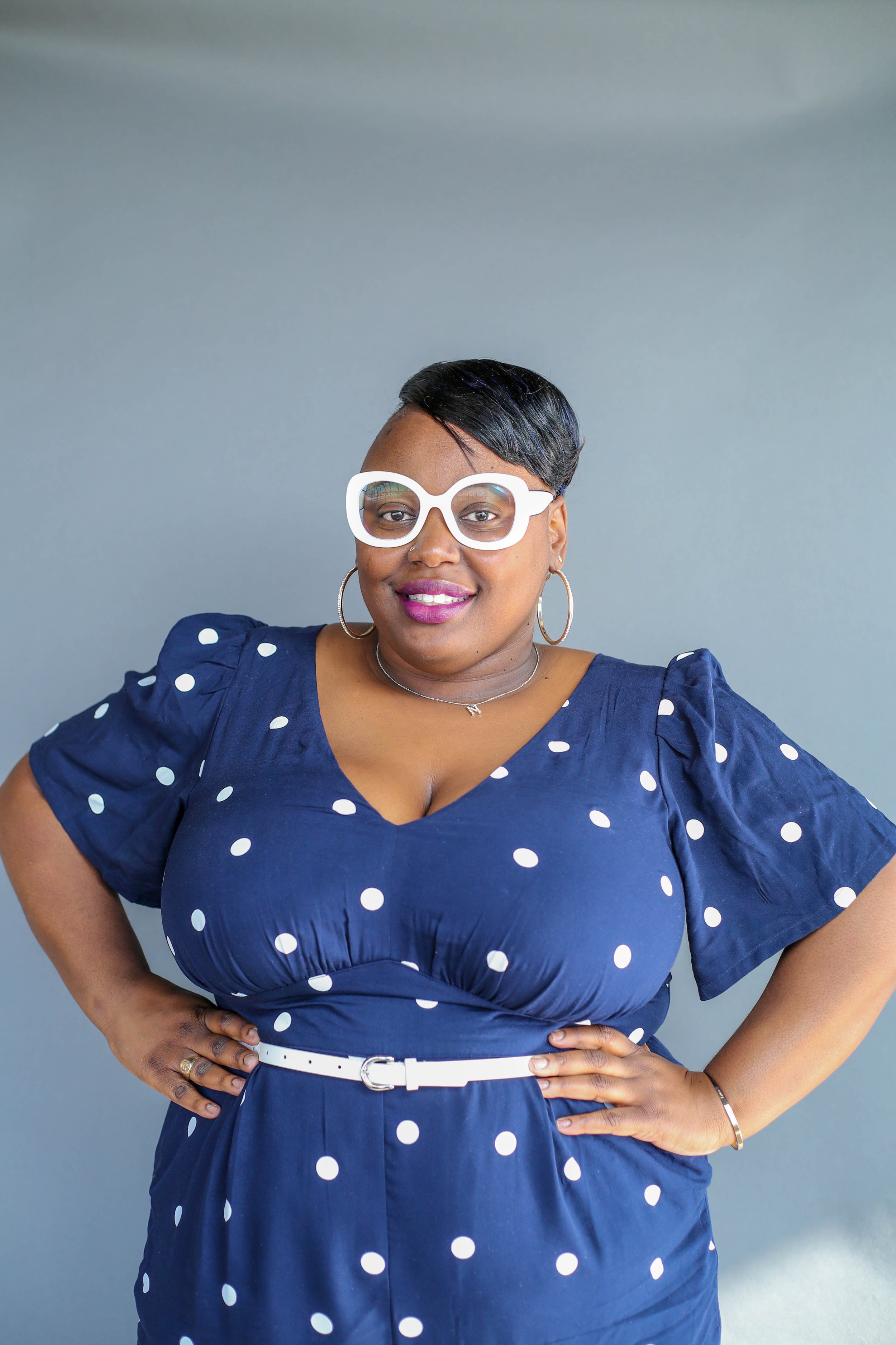 A woman with short black hair wearing white glasses, hoop earrings, a necklace, a white belt, and a navy blue dress with white polka dots, standing against a gray background.