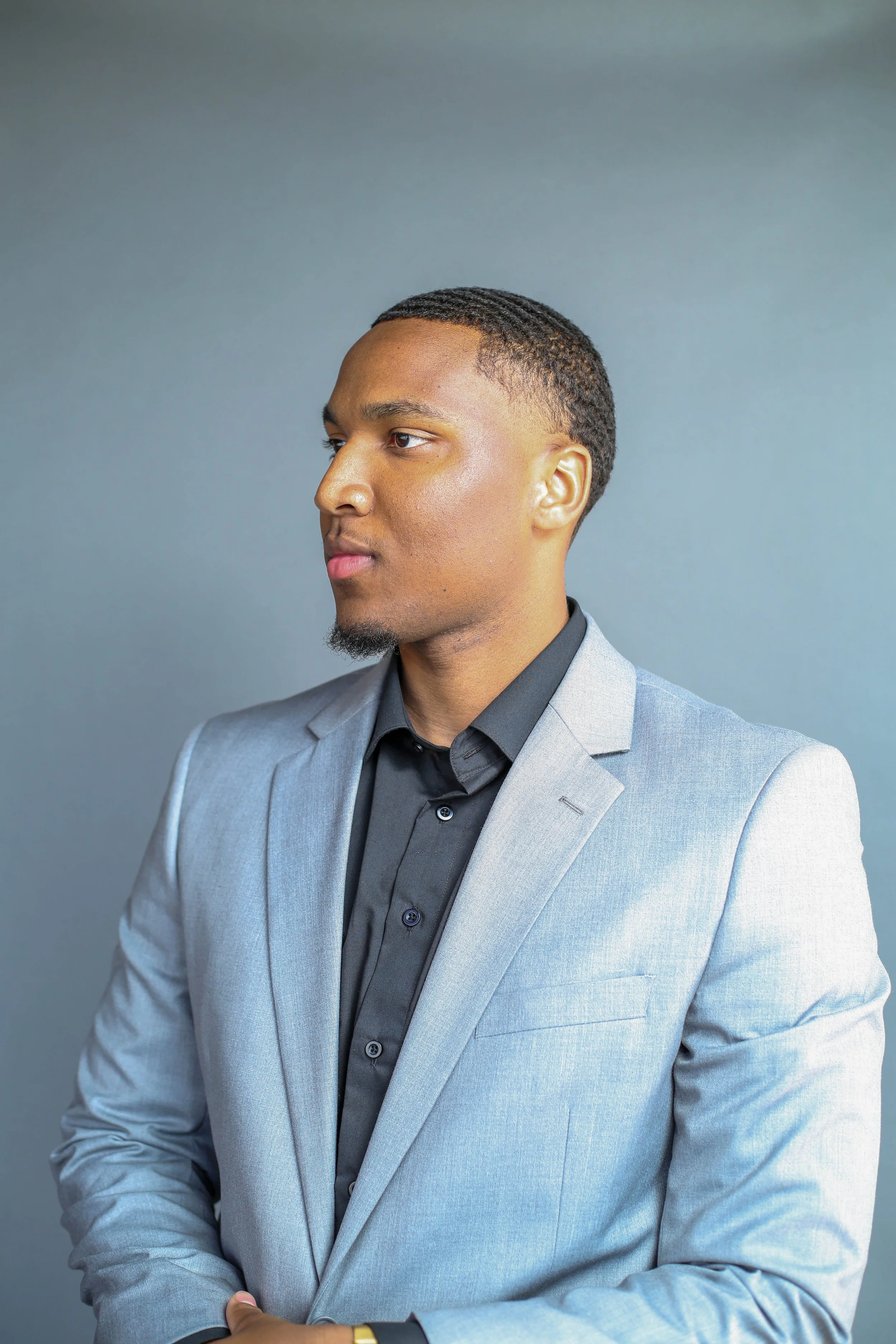 A young man in a light gray suit and dark gray shirt with a gold watch shows confidence with his arms crossed, standing against a plain gray background.