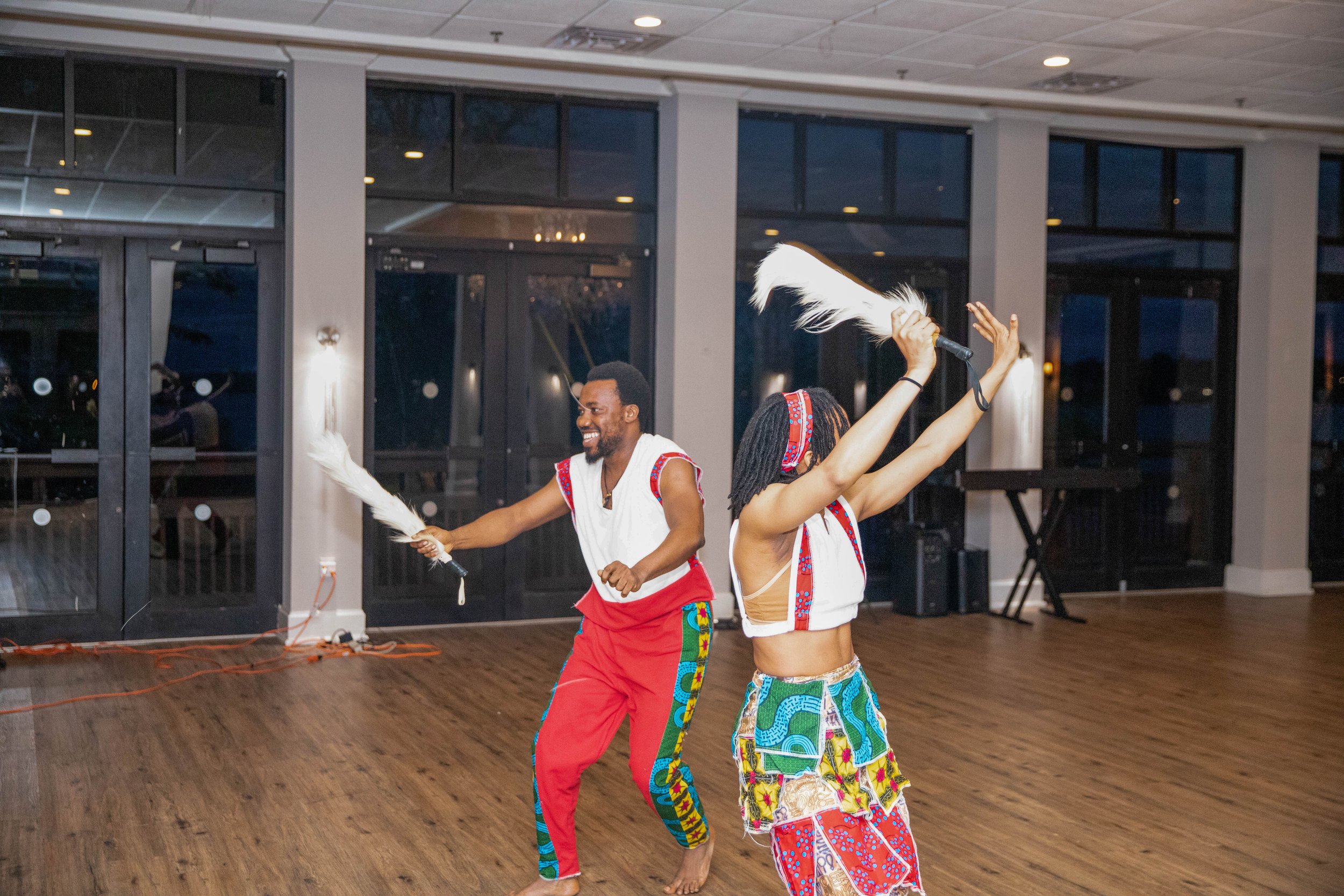 Two barefoot performers in colorful traditional African attire dancing with feathered fans in a modern indoor space with large windows and wooden flooring.