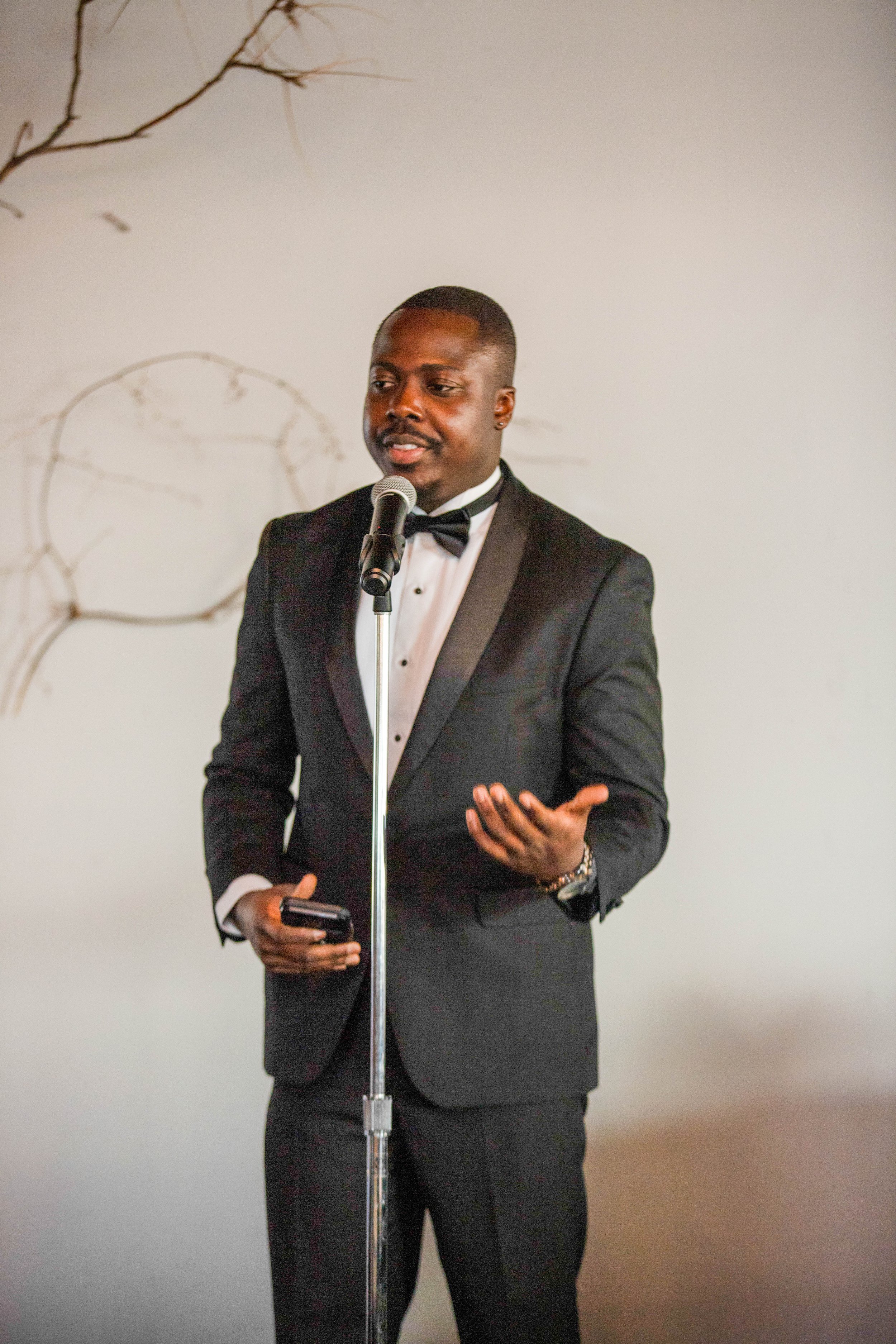 A man in a black tuxedo with a bow tie is giving a speech or presentation behind a microphone, holding a phone in his right hand and gesturing with his left hand.