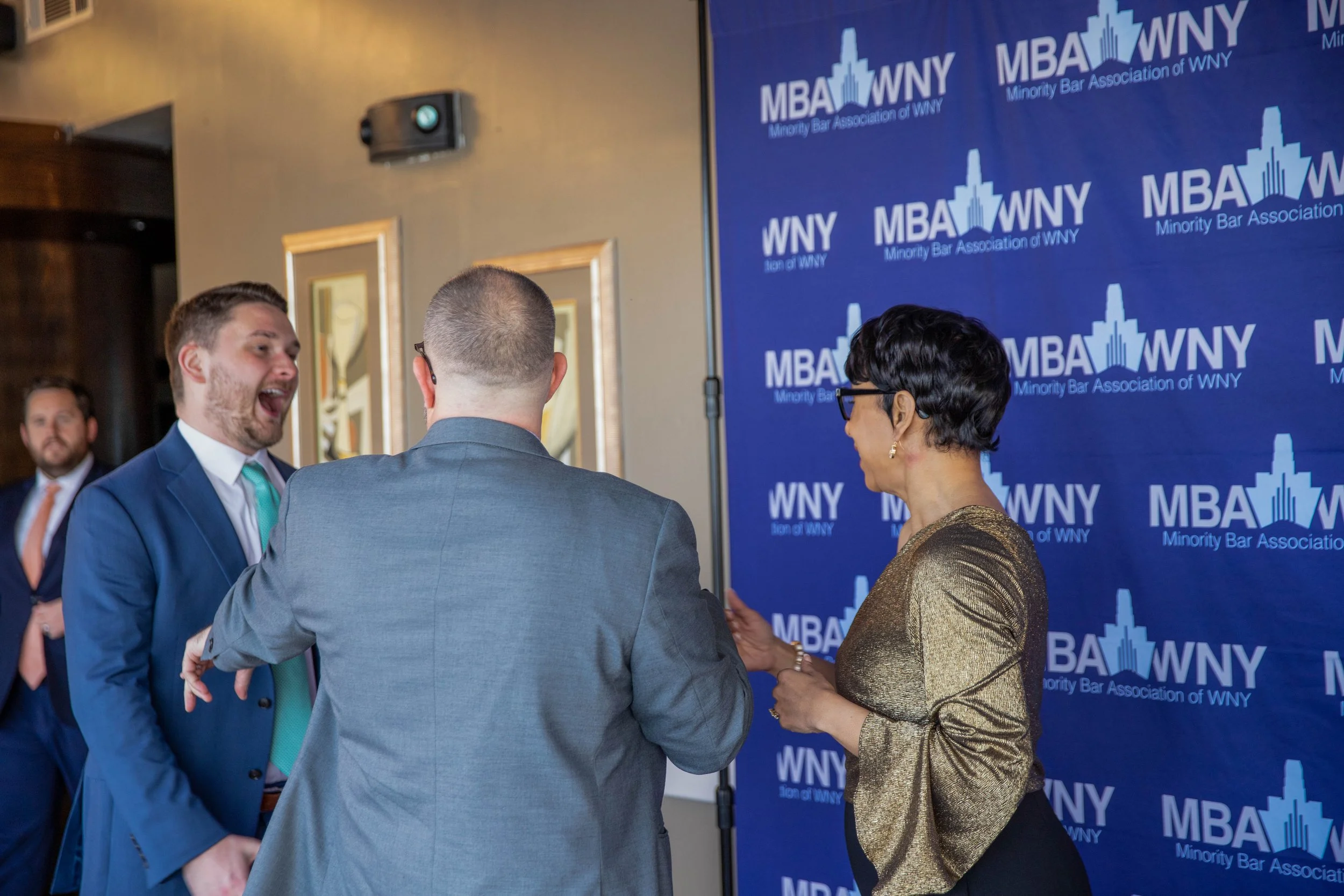 People talking at a professional event with a backdrop displaying the logo of the Minority Bar Association of WNY.