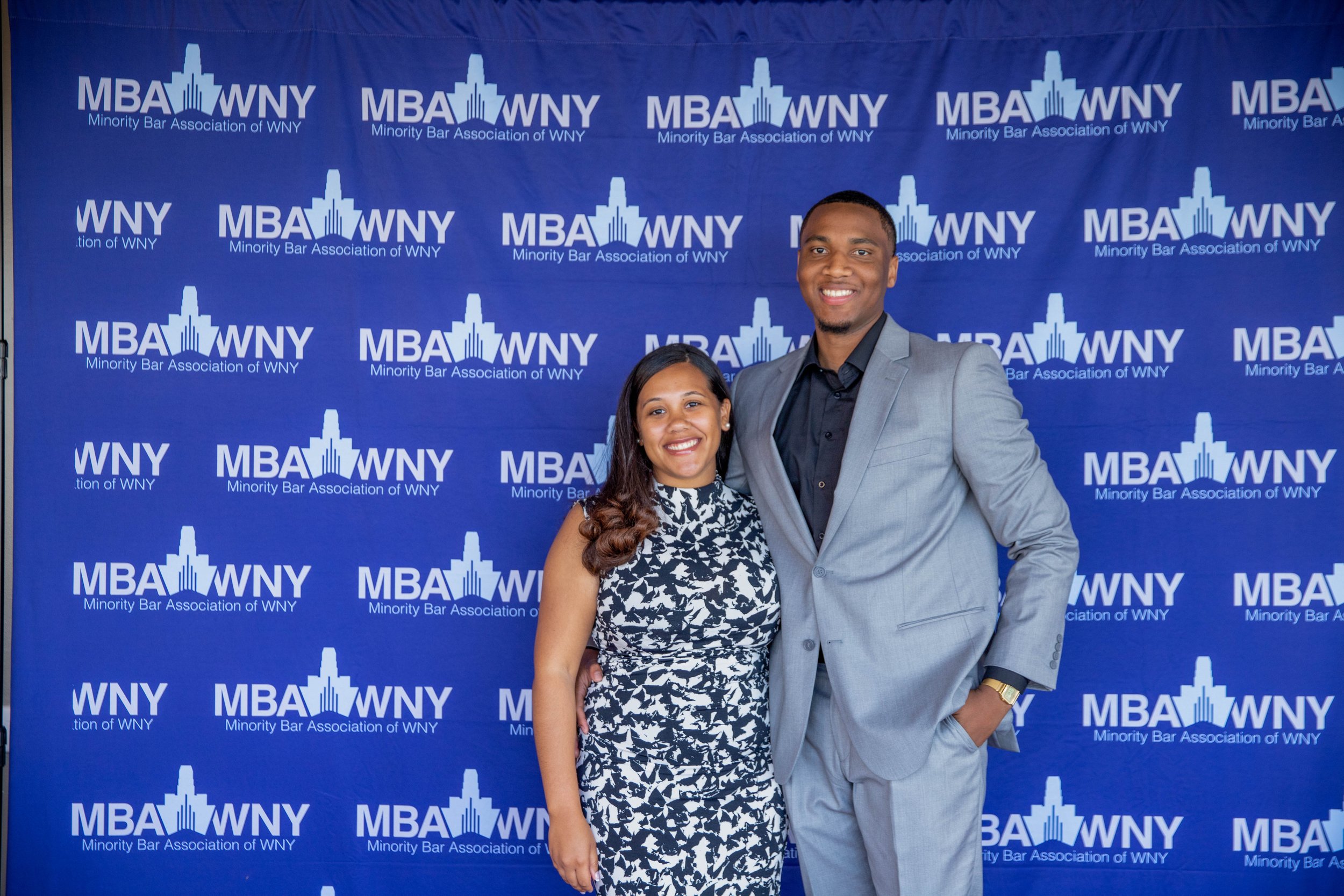 Two young adults, a woman and a man, standing in front of a blue backdrop with the logo and text of the Minority Bar Association of WNY. The woman is wearing a black and white patterned dress, and the man is dressed in a light gray suit with a black 