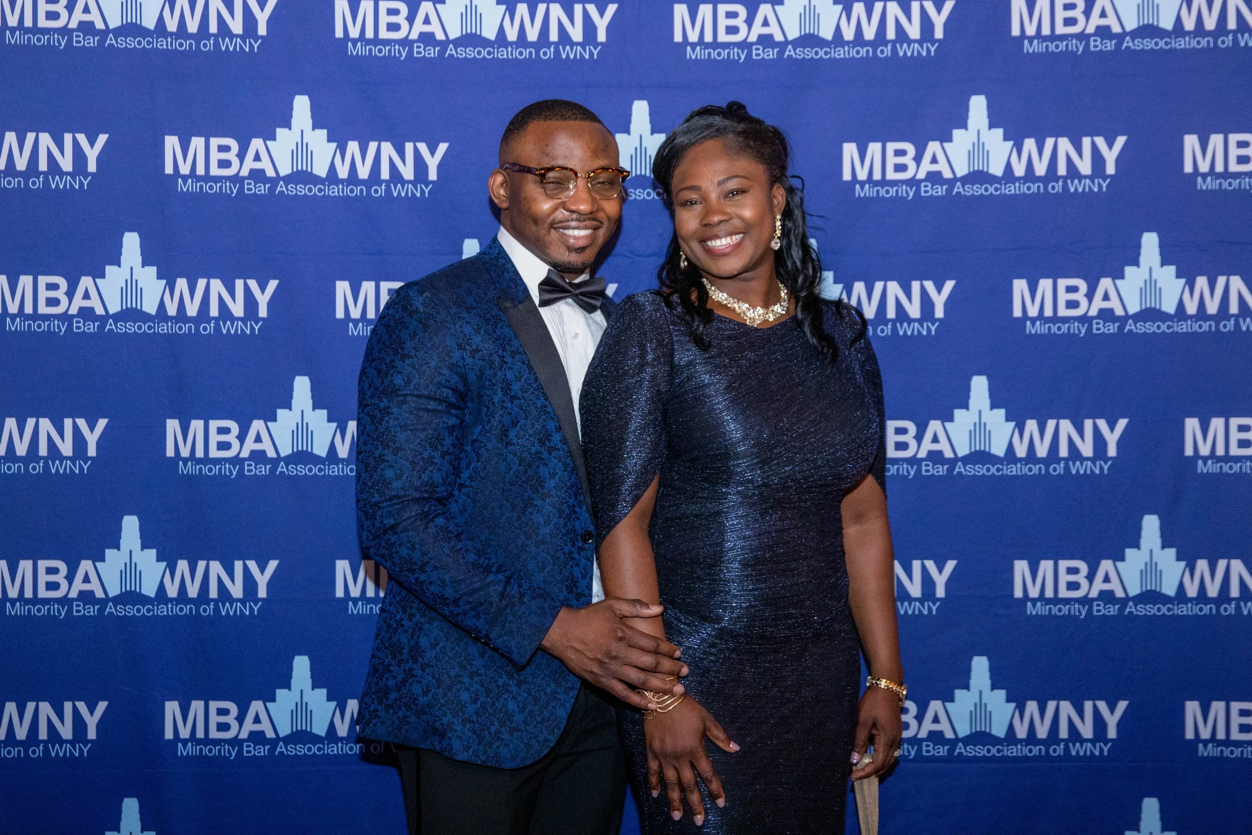 A man and a woman dressed in formal attire posing for a photo at an event with a blue backdrop displaying the logo and text for the Minority Bar Association of WNY, featuring a city skyline graphic.