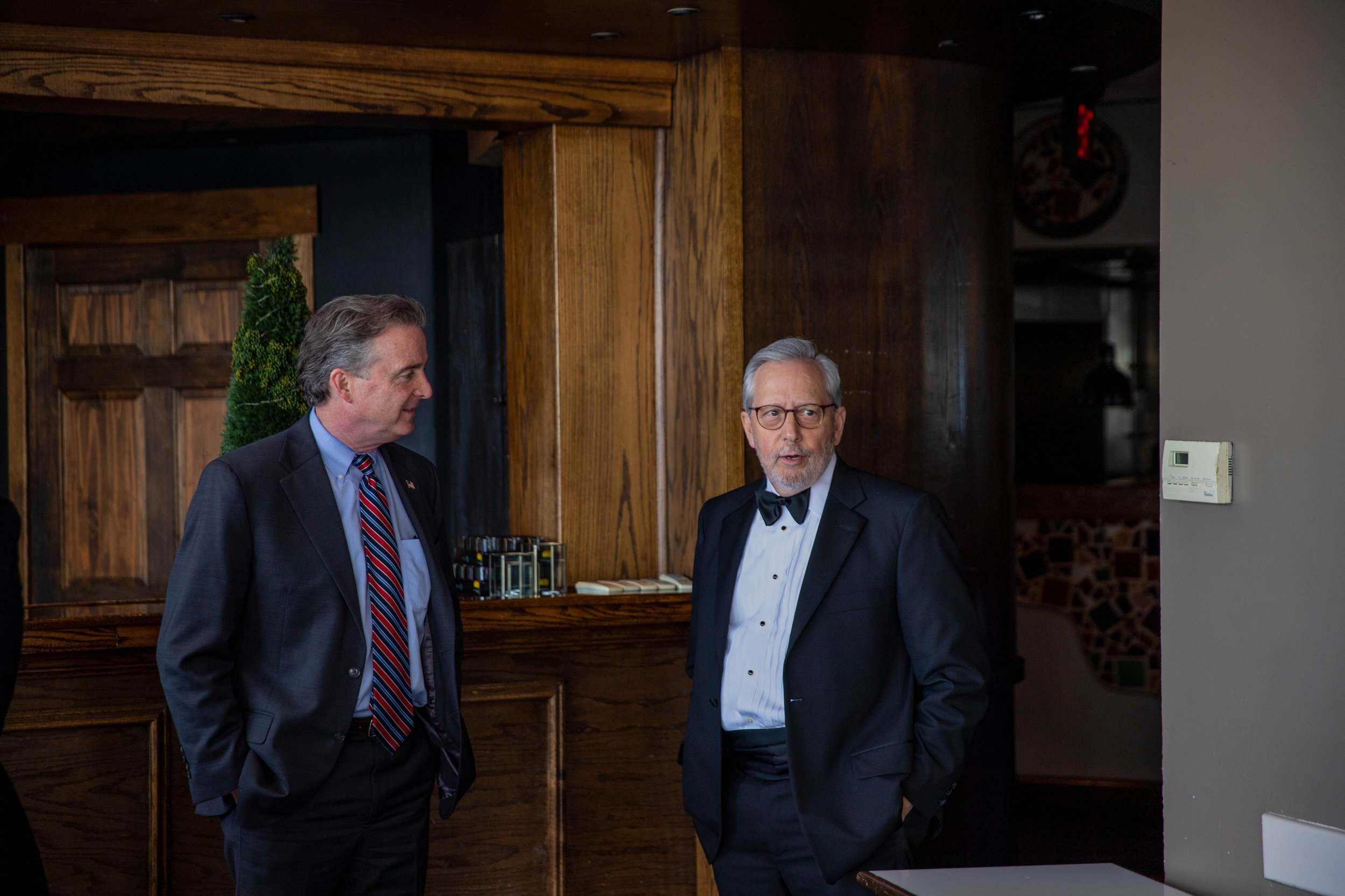 Two men in tuxedos engaging in conversation in a wood-paneled room, with a decorative plant and a dartboard in the background.