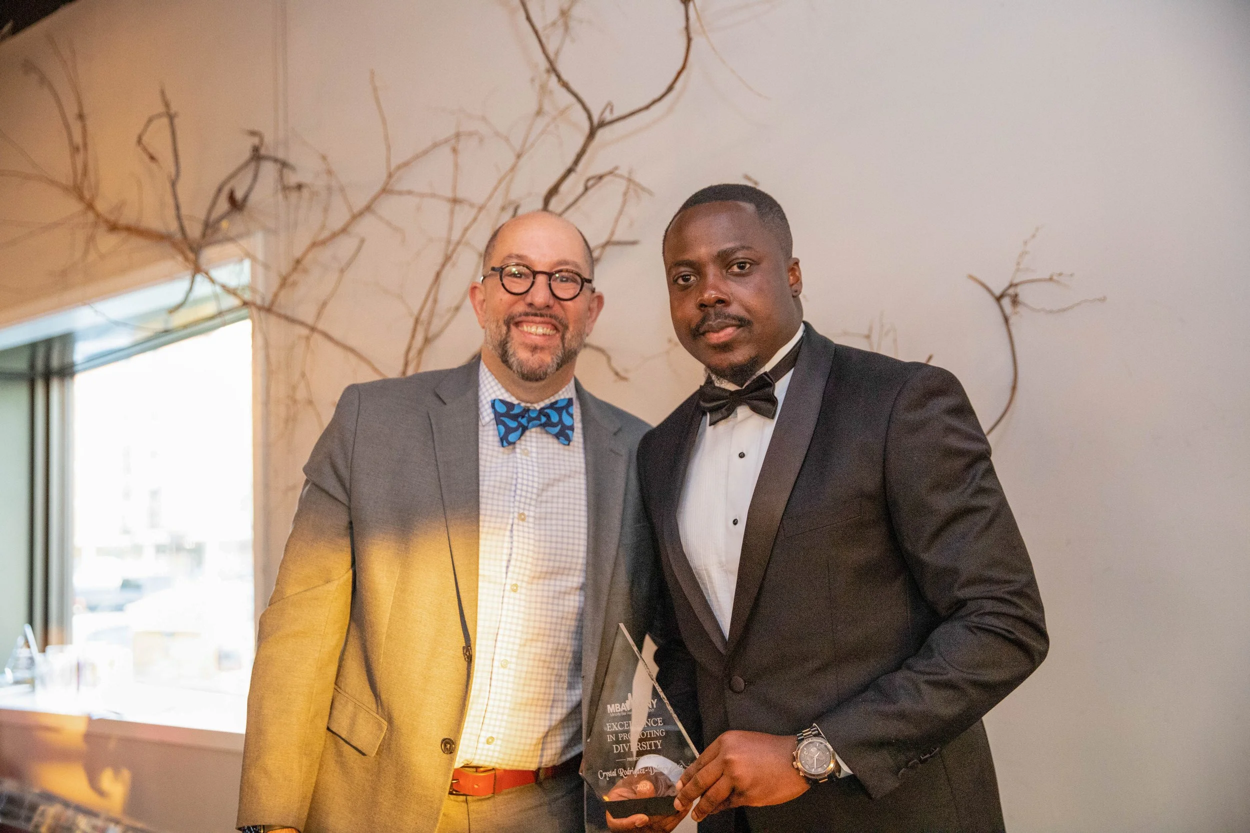 Two men in formal attire standing together indoors, one with glasses and a bow tie, the other holding an award, with a decorative wall behind them.