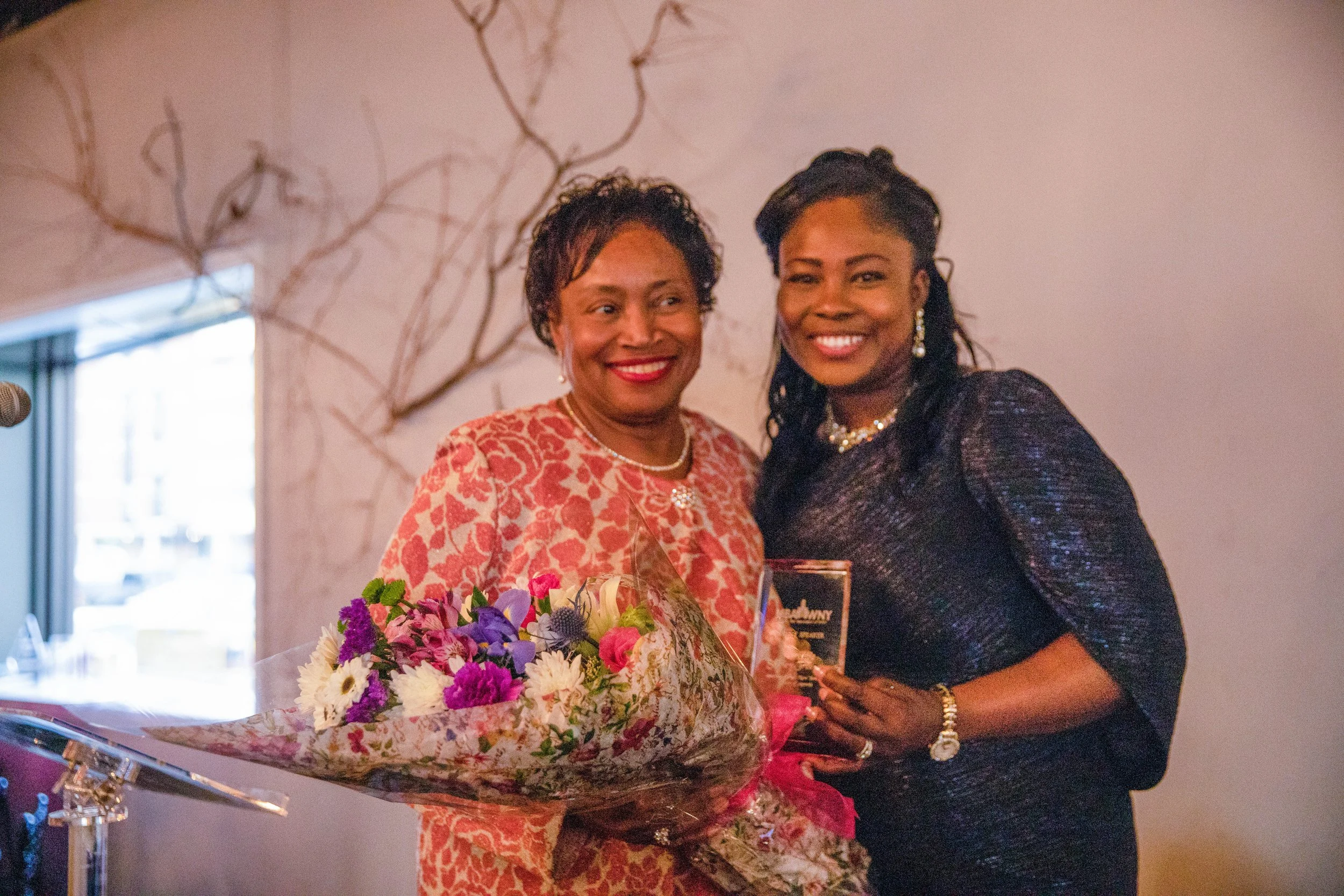 Two women smiling, one holding a bouquet of colorful flowers and the other holding a glass of champagne, at a celebratory event indoors.