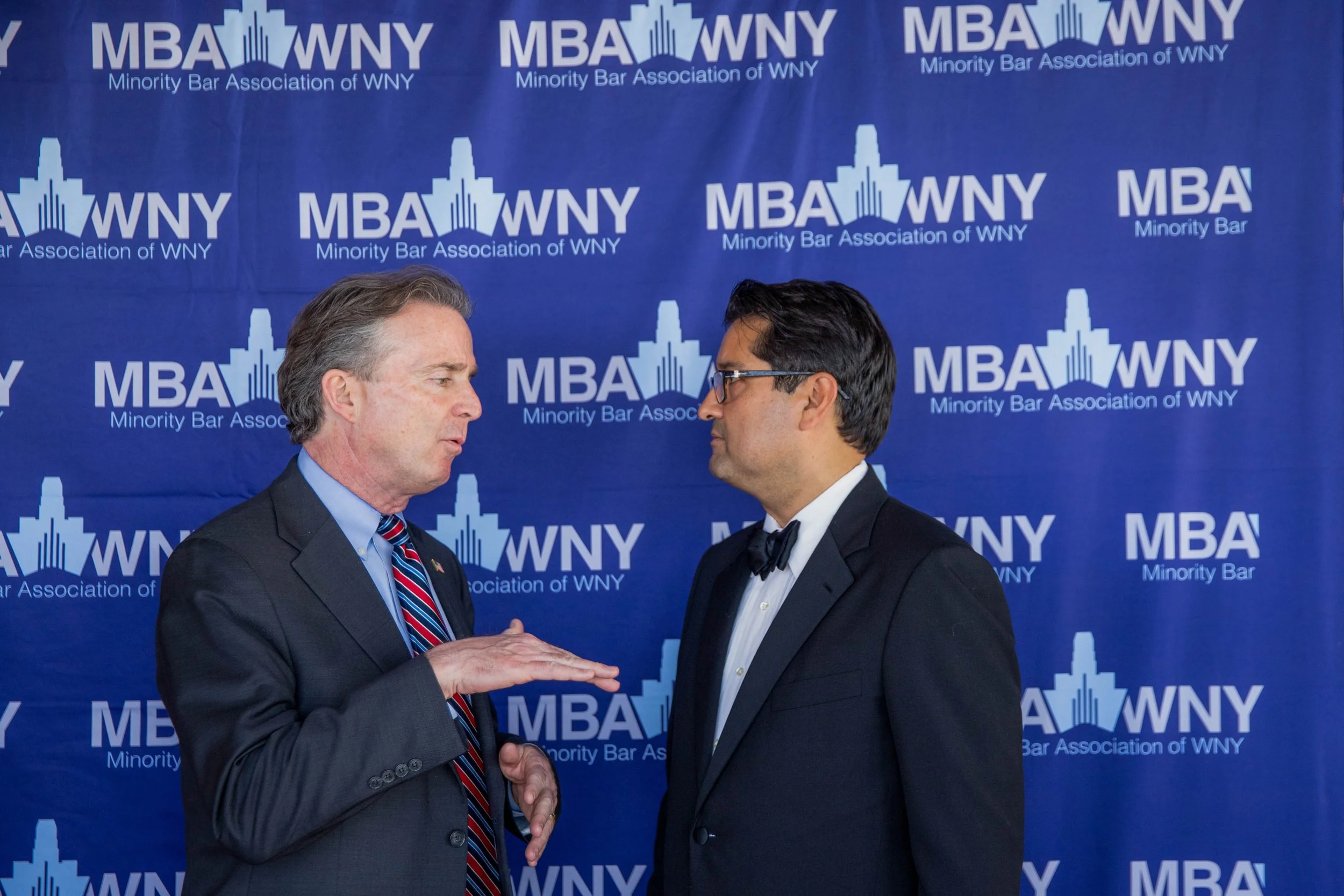 Two men in suits shaking hands and facing each other in front of a blue background with the text read 'MBA WNY Minority Bar Association of WNY' and a logo of a city skyline.