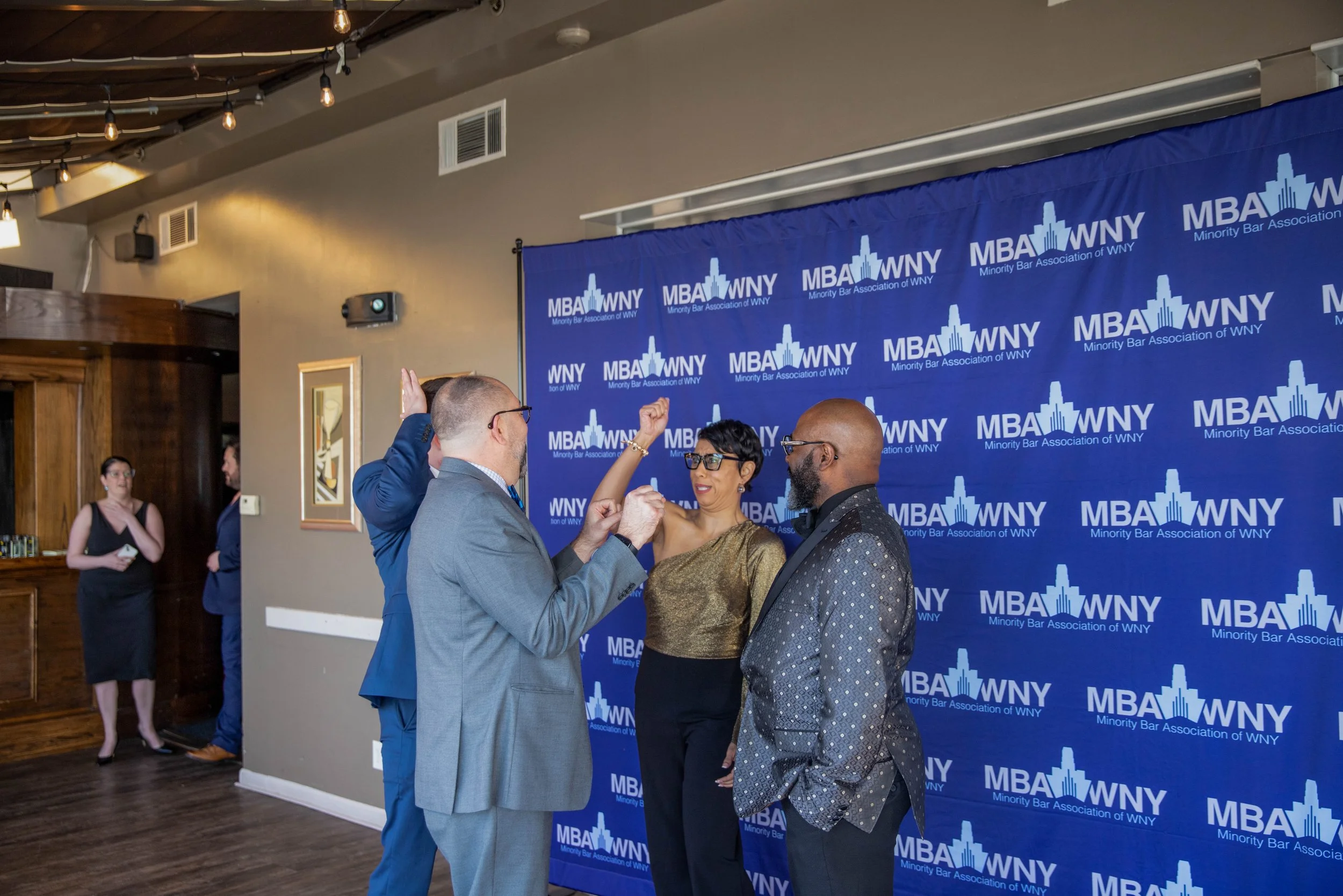 Four people are standing in front of a blue backdrop with the logo and name of the Minority Bar Association of WNY. Two men appear to be engaged in conversation, one of whom is raising a hand, while a woman looks on. A woman in a black dress and glas
