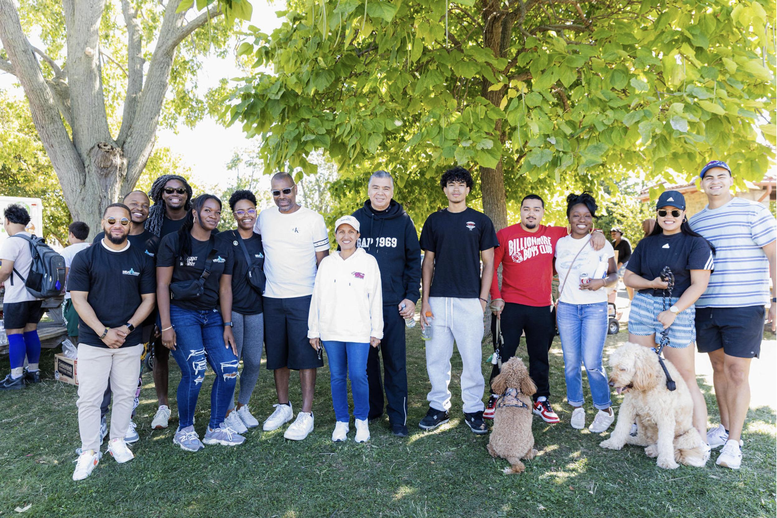 A diverse group of people standing outdoors under a large green tree, smiling at the camera, with some dogs and others with backpacks.