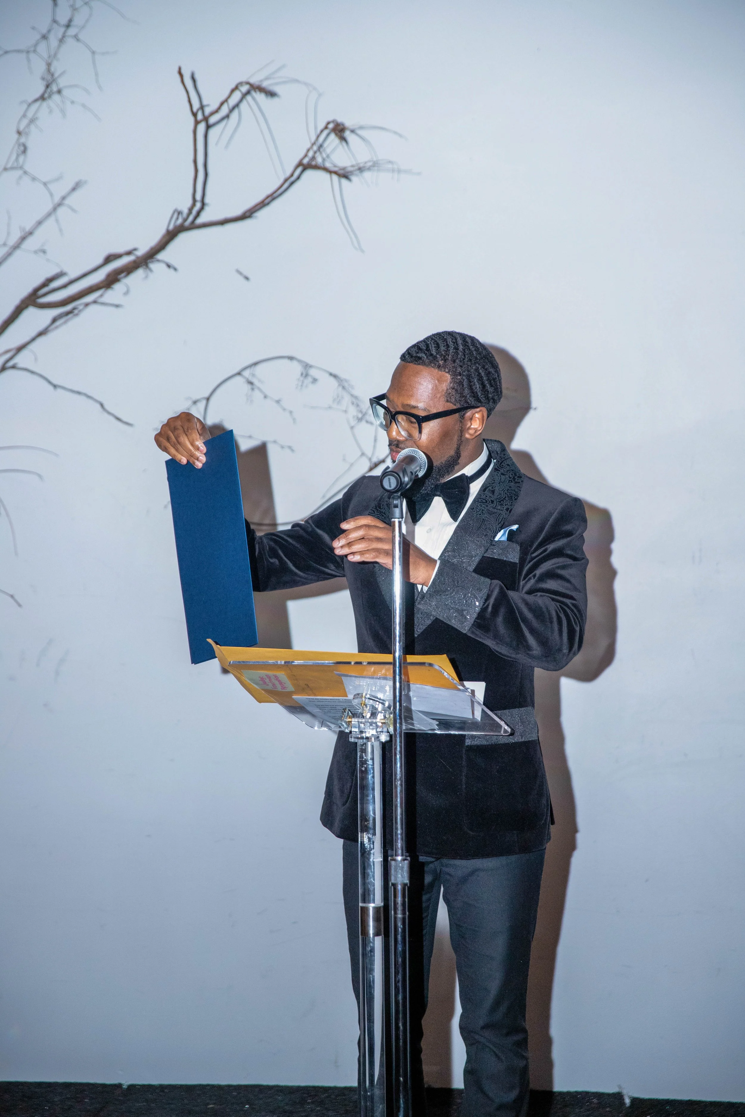 A man in a tuxedo and glasses stands at a clear acrylic podium with a microphone, holding a dark blue folder. Behind him is a plain white wall adorned with a few bare tree branches.