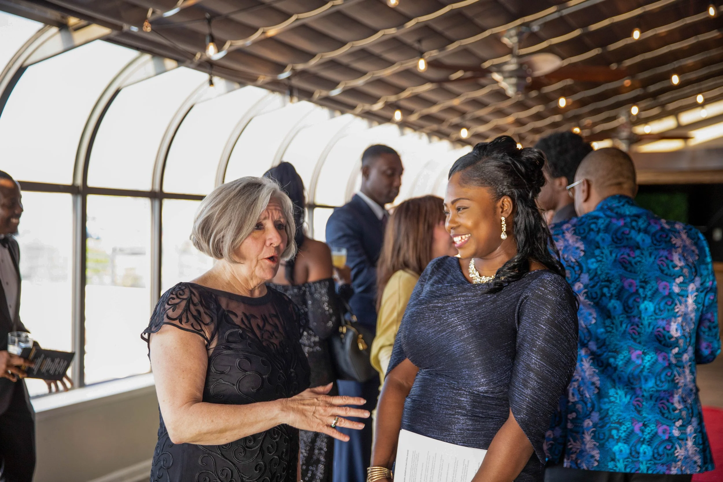 Two women, one with gray hair and a black lace dress, and the other with black hair and a dark blue dress, are engaging in conversation at a social event. The background shows other people talking and mingling in a well-lit indoor space with large wi