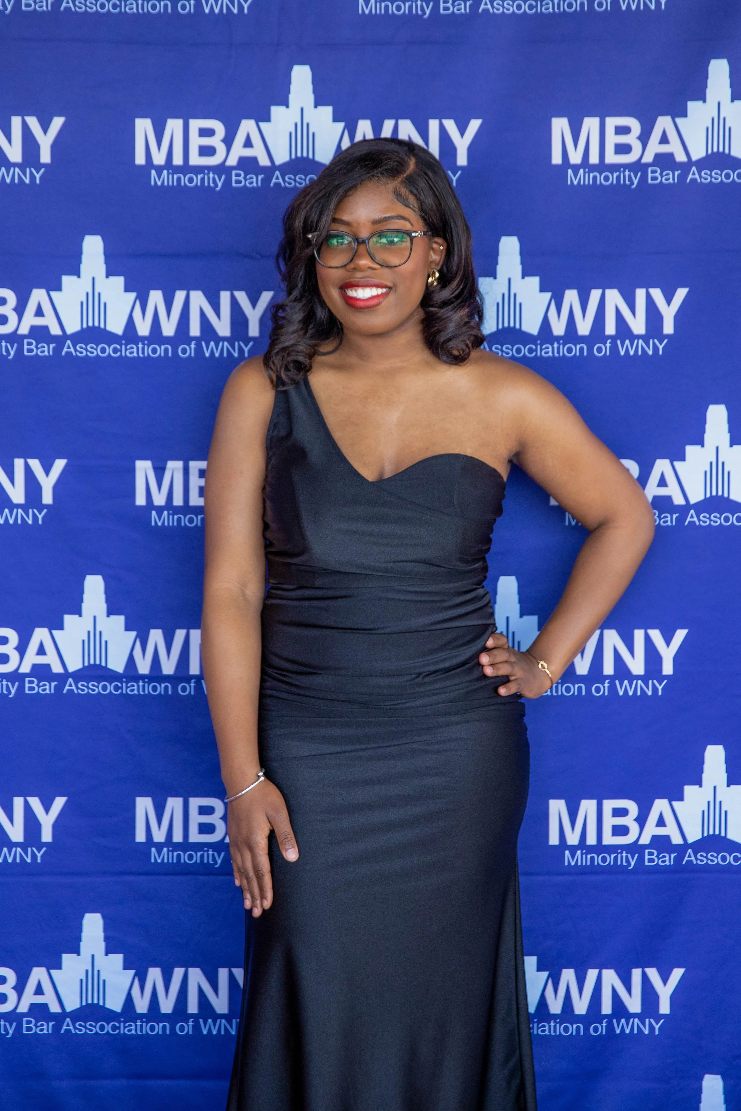 A young woman with shoulder-length curly black hair, wearing glasses, a black one-shoulder dress, standing in front of a blue backdrop with the logo and text of the Minority Bar Association of WNY.