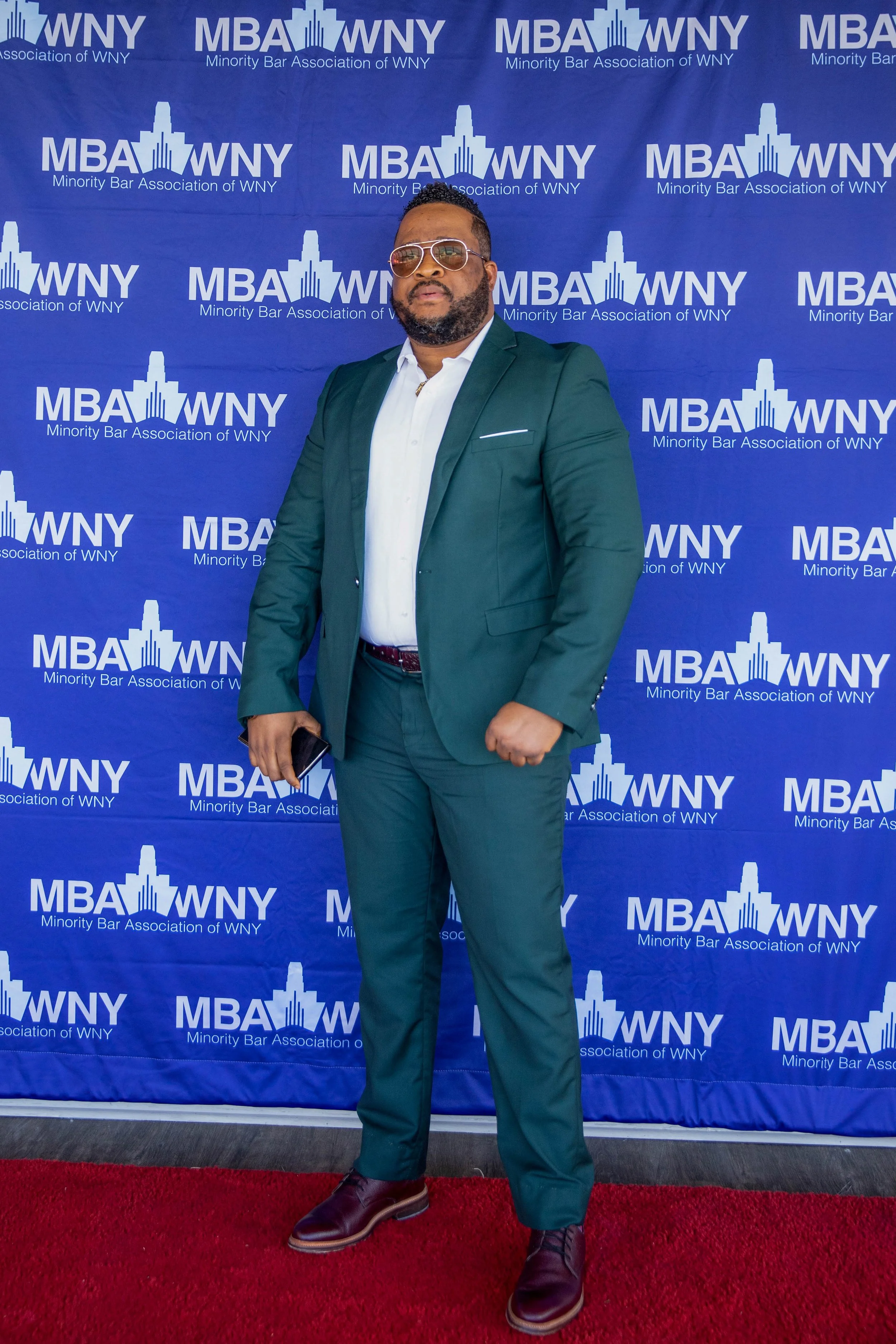 A man in a teal suit and white shirt standing on a red carpet in front of a blue backdrop with logos for the Minority Bar Association of WNY. He is wearing sunglasses and holding a phone in his right hand.
