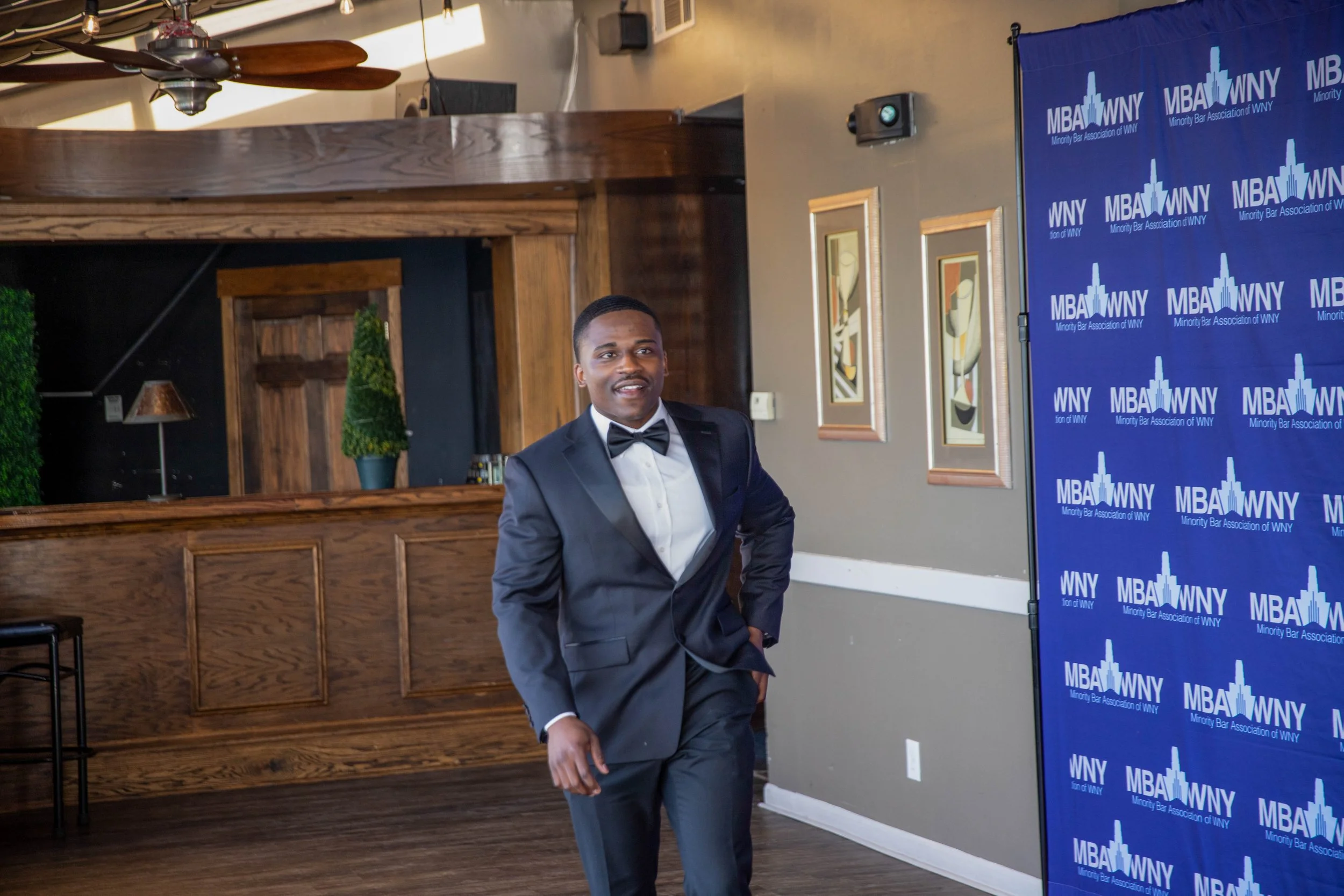 A man in a tuxedo with a bow tie standing indoors at an event. Behind him is a blue banner with white text that reads "MBA WNY" and "Minority Bar Association of WNY." The room has wooden panels, framed artwork, and potted plants. A ceiling fan is vis