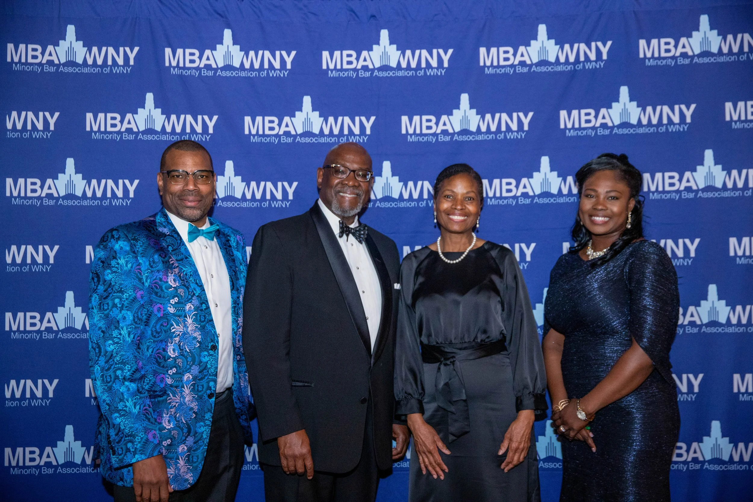 Group of four people attending an event at the Minority Bar Association of WNY, standing in front of a blue backdrop with the organization's logo.