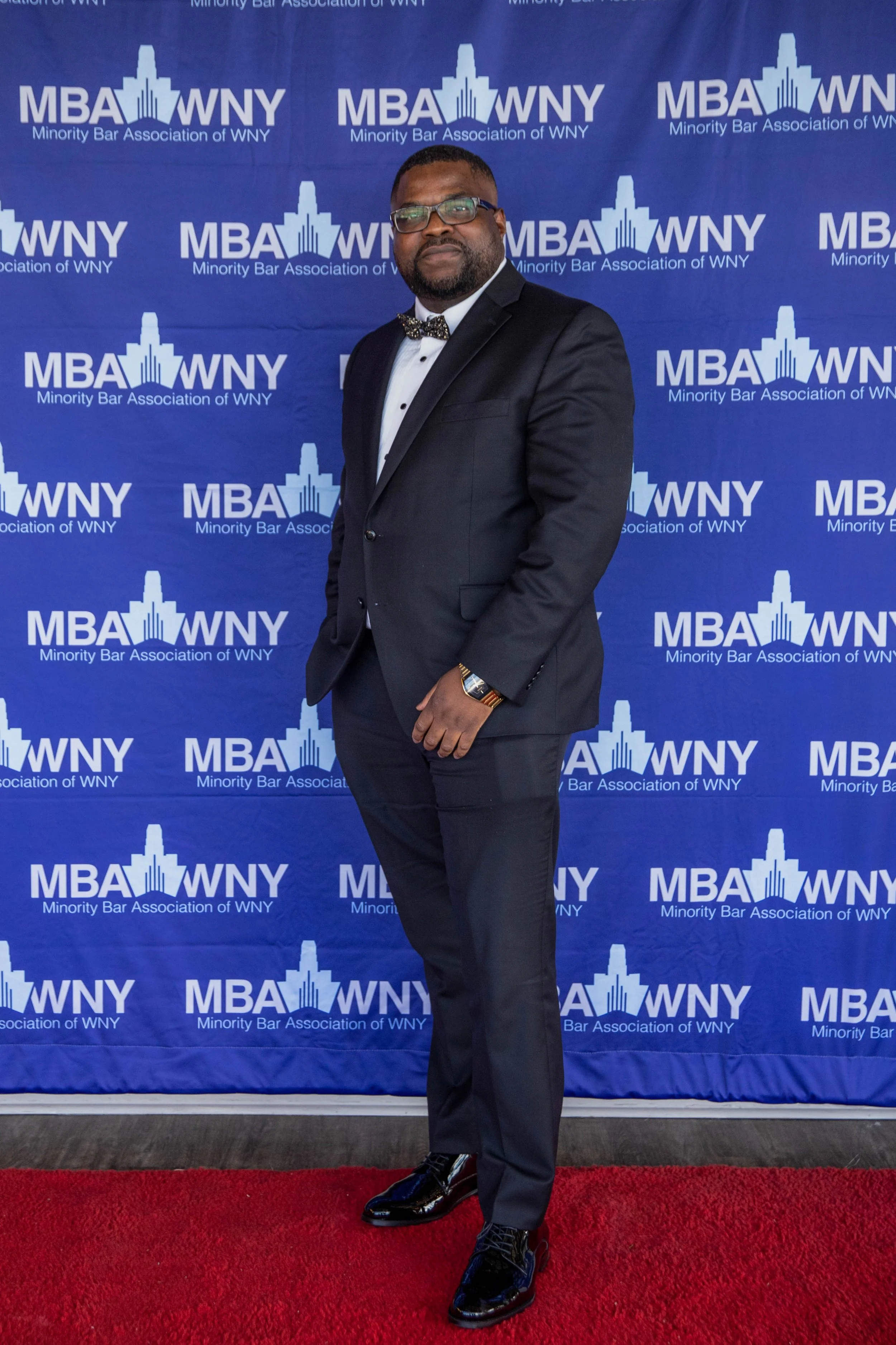 A man in a tuxedo with a bow tie and glasses standing on a red carpet in front of a blue backdrop with the repeated logo of the Minority Bar Association of WNY, which features a stylized building and the acronym MBA WNY.