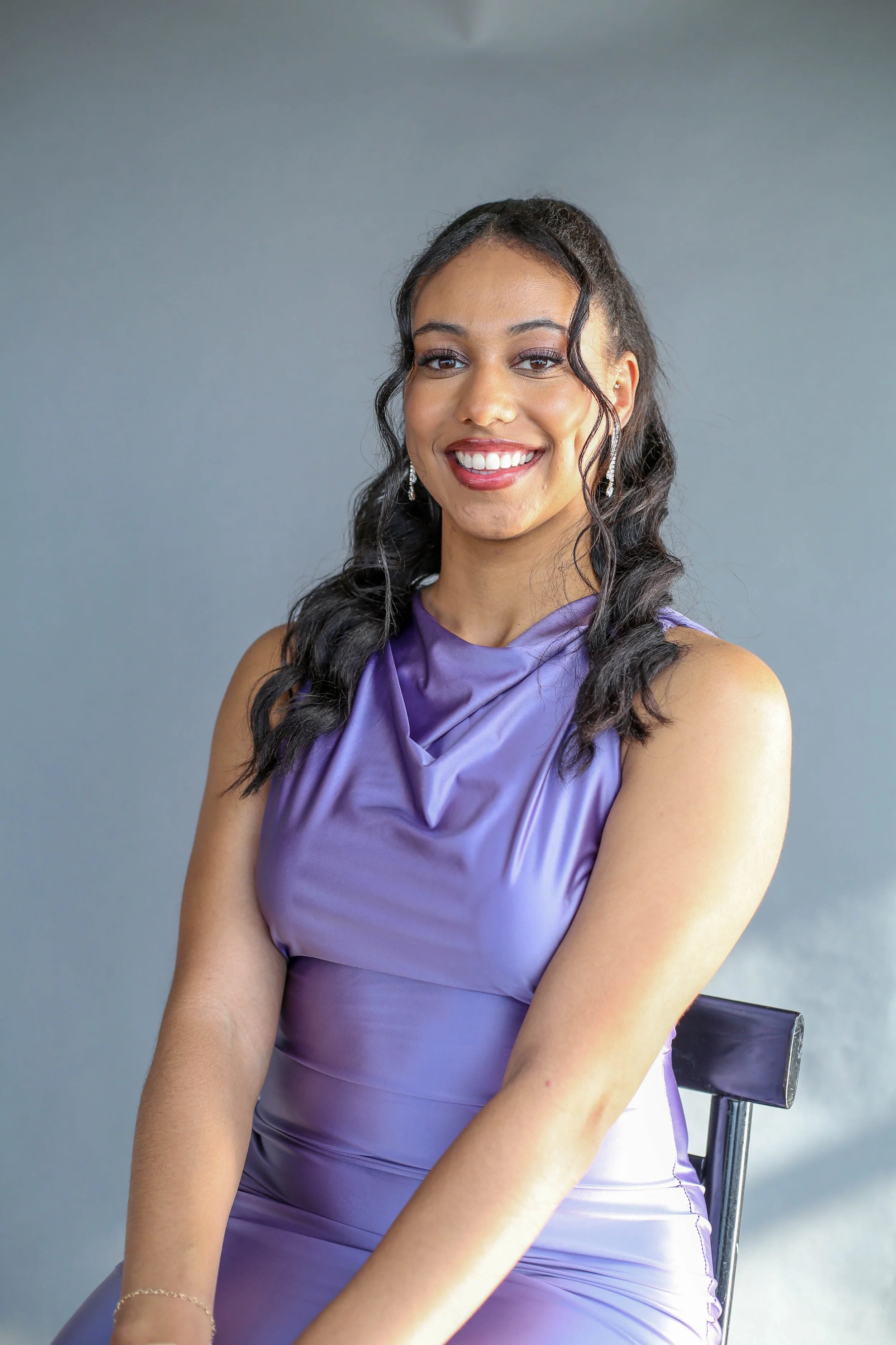 A woman in a purple satin dress sitting on a chair, smiling at the camera with a plain gray background.