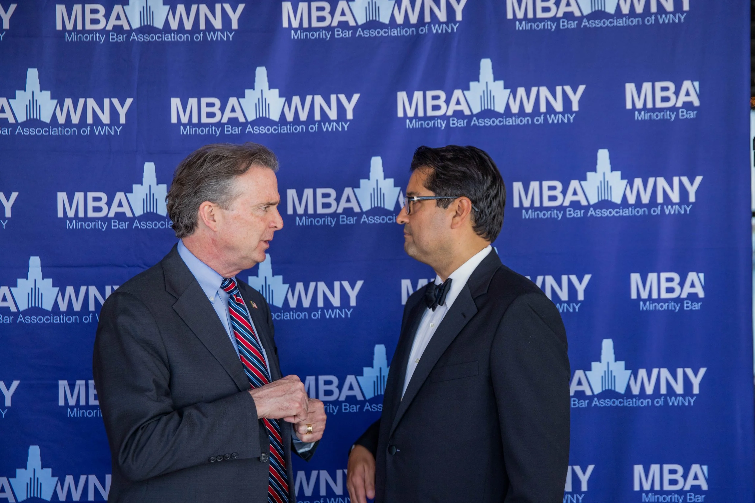 Two men in suits shaking hands at a formal event, standing in front of a blue backdrop with the logo and text of the Minority Bar Association of WNY.