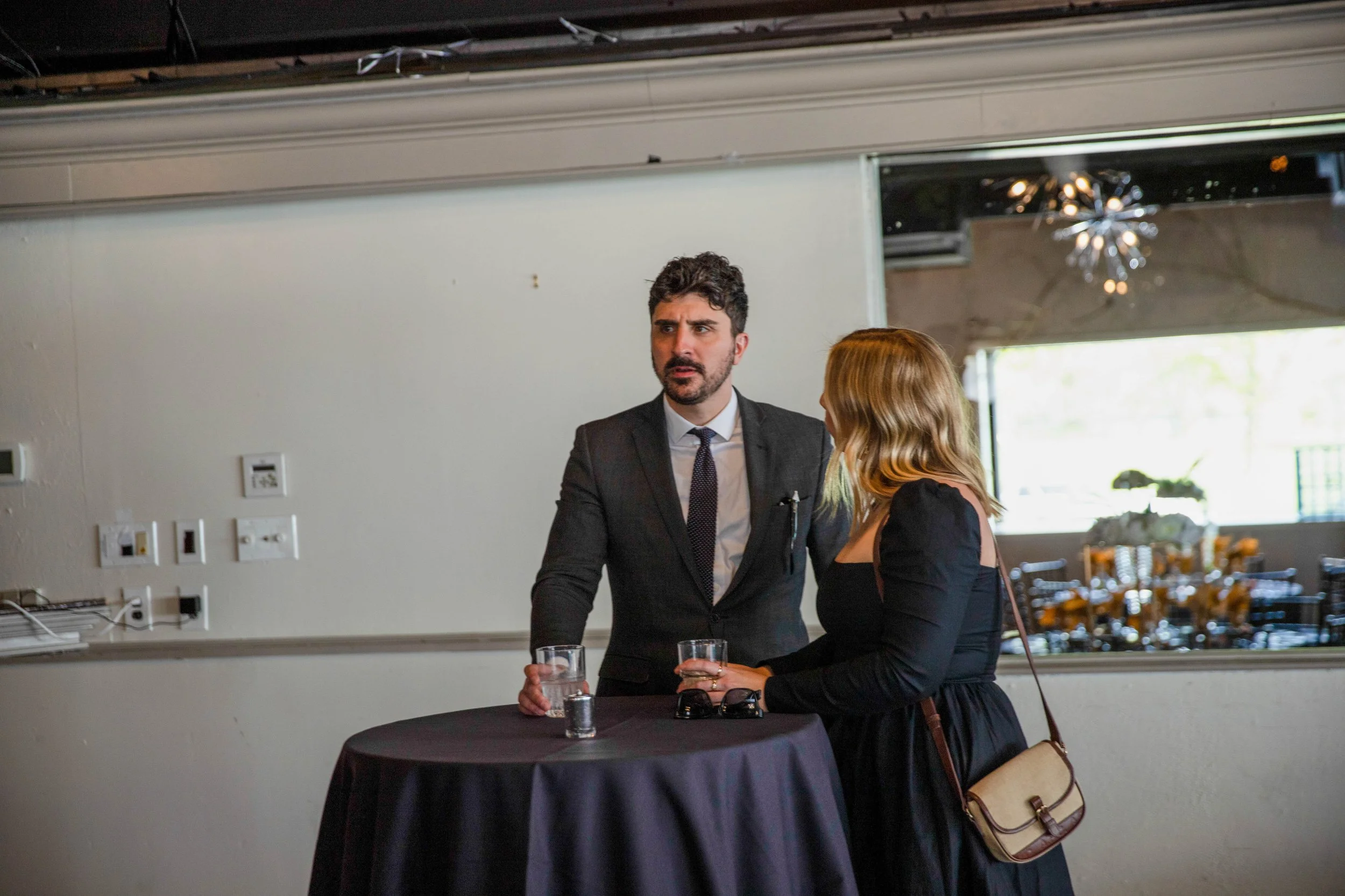 A man in a dark suit talking to a woman at a social event. They are standing near a table with glasses of water, with another woman partially visible. The setting appears to be indoors with modern decor.