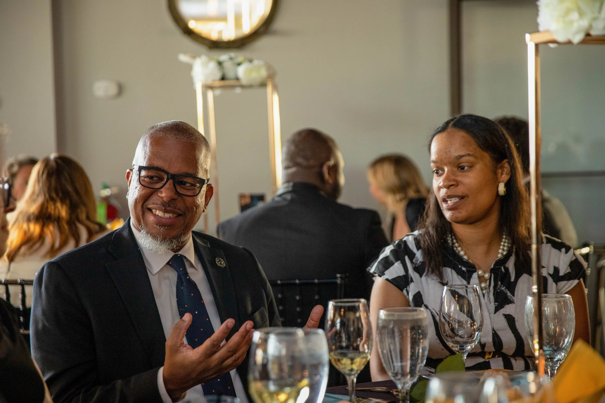 A man wearing glasses, a suit, and a tie is smiling and making a peace sign with his hand, sitting at a dinner table with glasses of white wine. Next to him, a woman wearing a pearl necklace and a black and white patterned dress is speaking. There ar