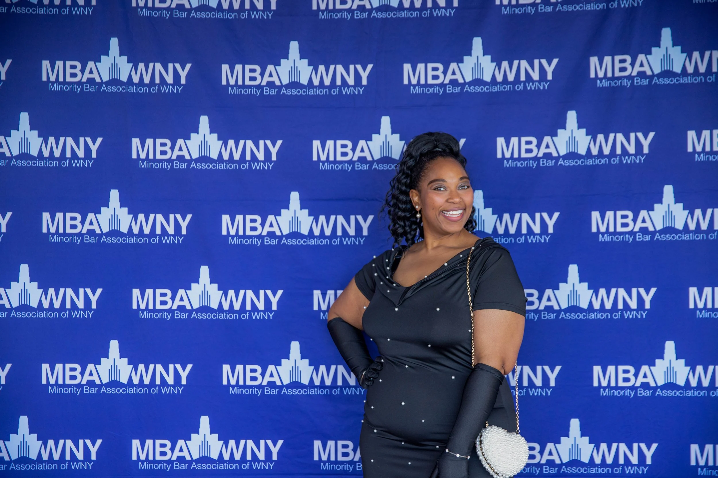 A woman smiling and posing in front of a backdrop with the logo of the Minority Bar Association of WNY, featuring a cityscape and the acronym MBA WNY.