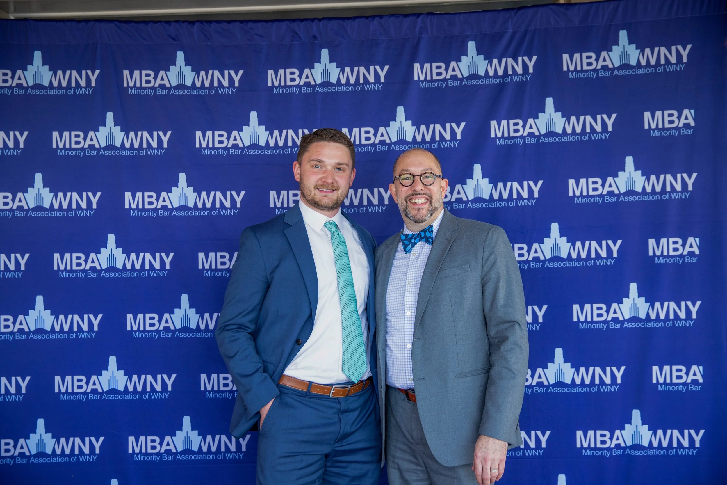 Two men standing in front of a blue backdrop with the logo and text of the Minority Bar Association of WNY. Both men are dressed in suits; one is wearing a light blue tie and the other has a blue bow tie with a pattern, and glasses on one of them. Th