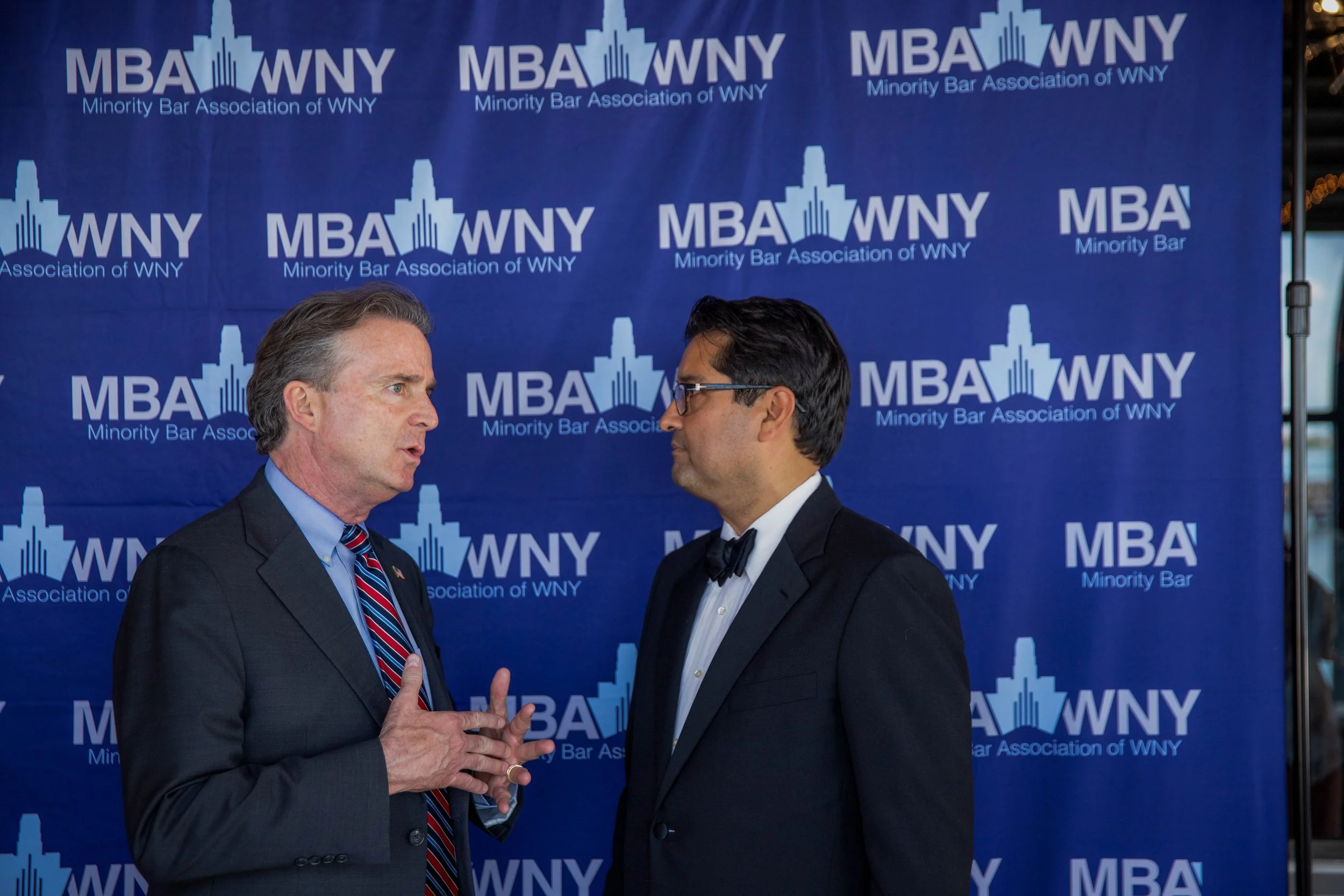 Two men in formal attire engaged in conversation with a blue backdrop behind them that has the logo of the Minority Bar Association of WNY repeated across it.