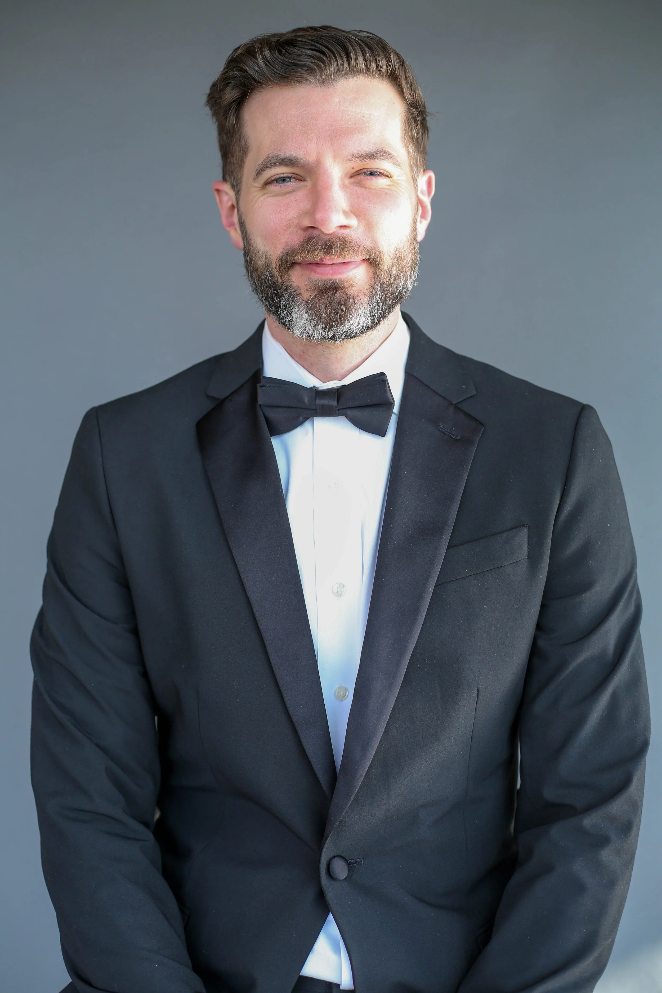 A man wearing a black tuxedo with a bow tie, white shirt, and short brown hair with a beard and mustache, is smiling gently against a plain gray background.