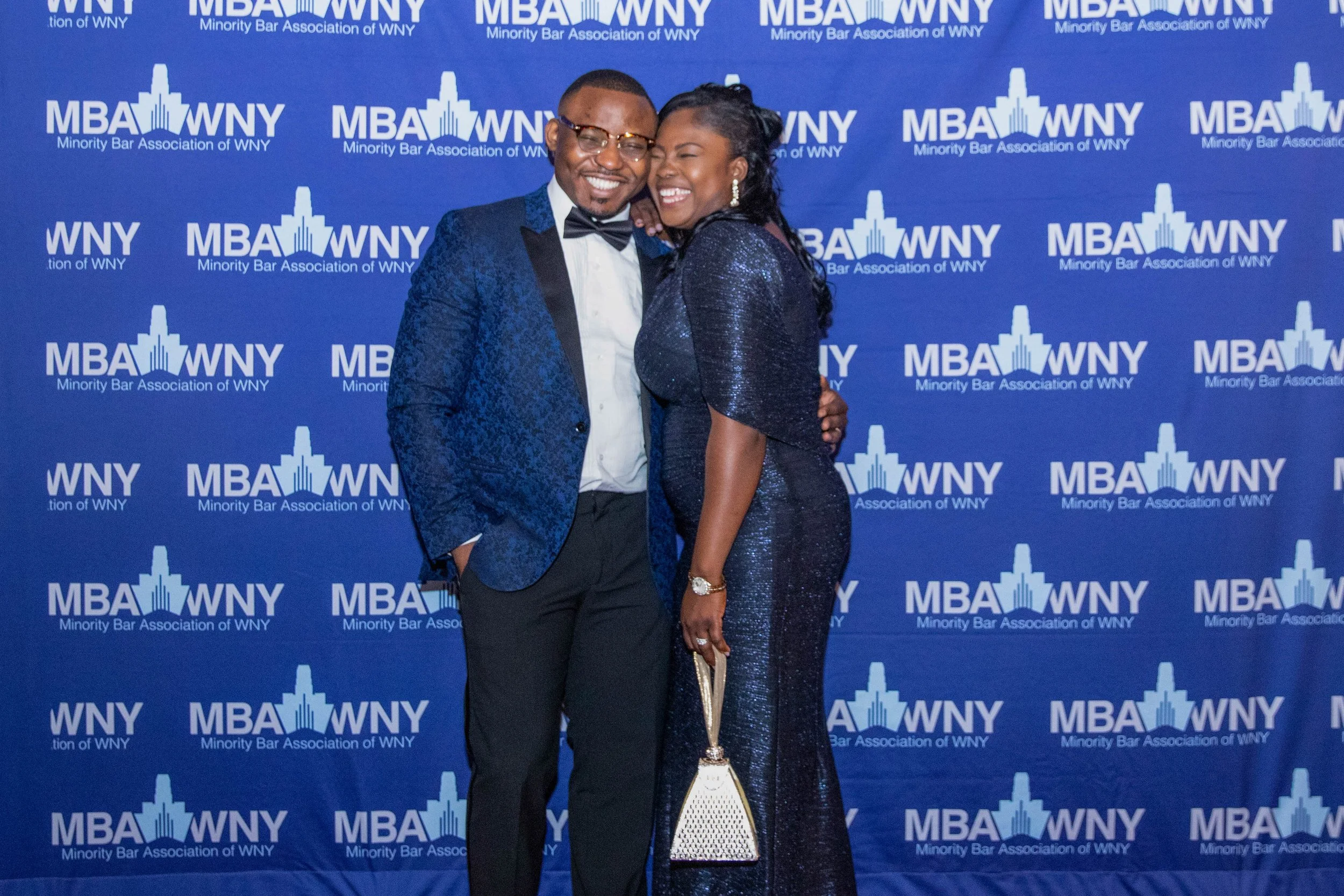 Two people standing in front of a blue backdrop with the text 'MBA WNY Minority Bar Association of WNY'. They are dressed formally, smiling, and embracing each other. The person on the left is wearing glasses and a blue tuxedo. The person on the righ