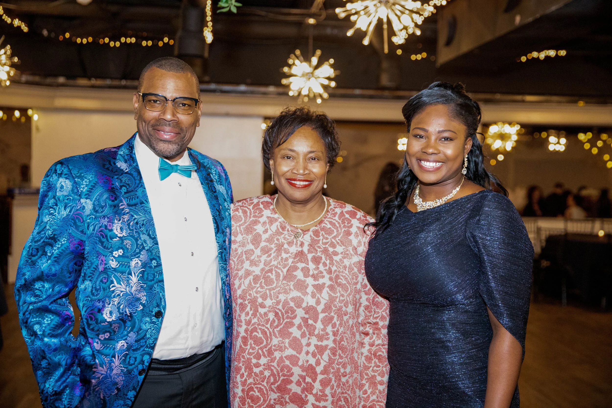 Three people standing together at an event, smiling for the camera. The man on the left is wearing a blue patterned blazer with a white shirt and teal bow tie, the woman in the middle is wearing a pink and white patterned dress, and the woman on the 