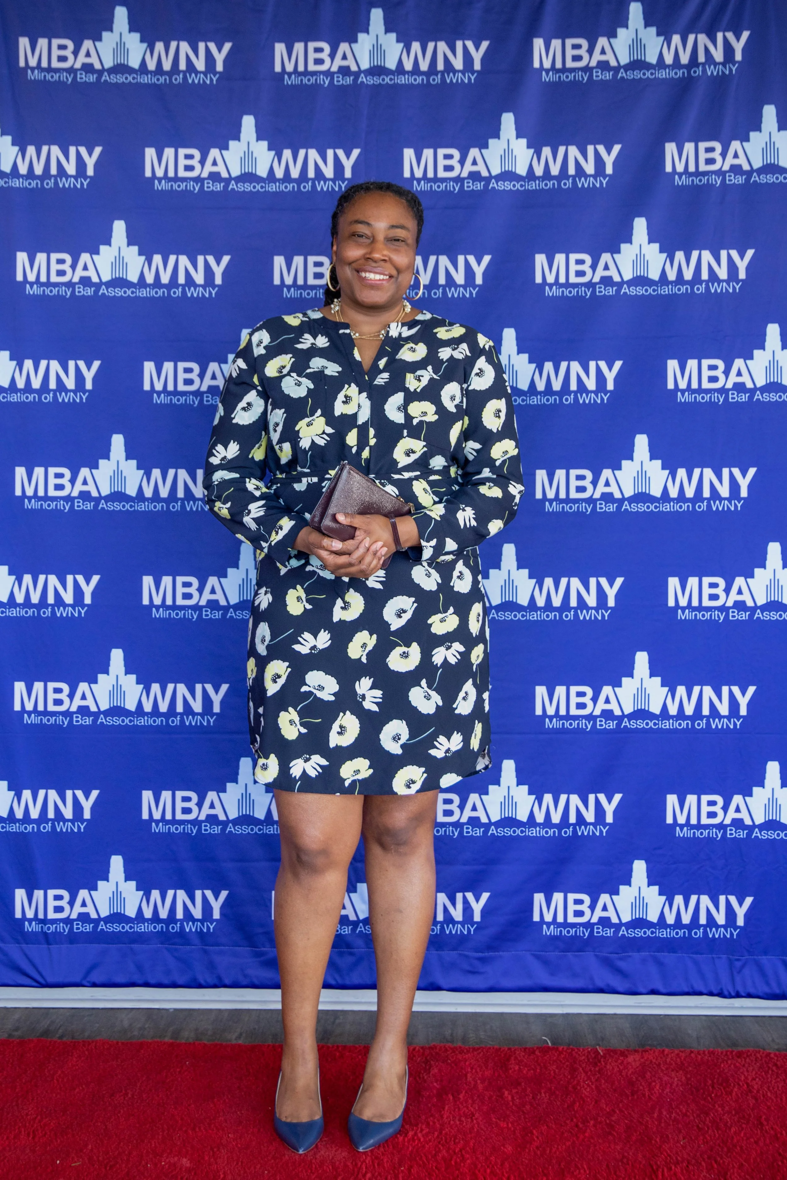 Woman standing on red carpet in front of blue backdrop with white logos reading 'MBA WNY Minority Bar Association of WNY,' wearing a blue floral dress, blue high heels, and holding a small brown clutch, smiling at camera.