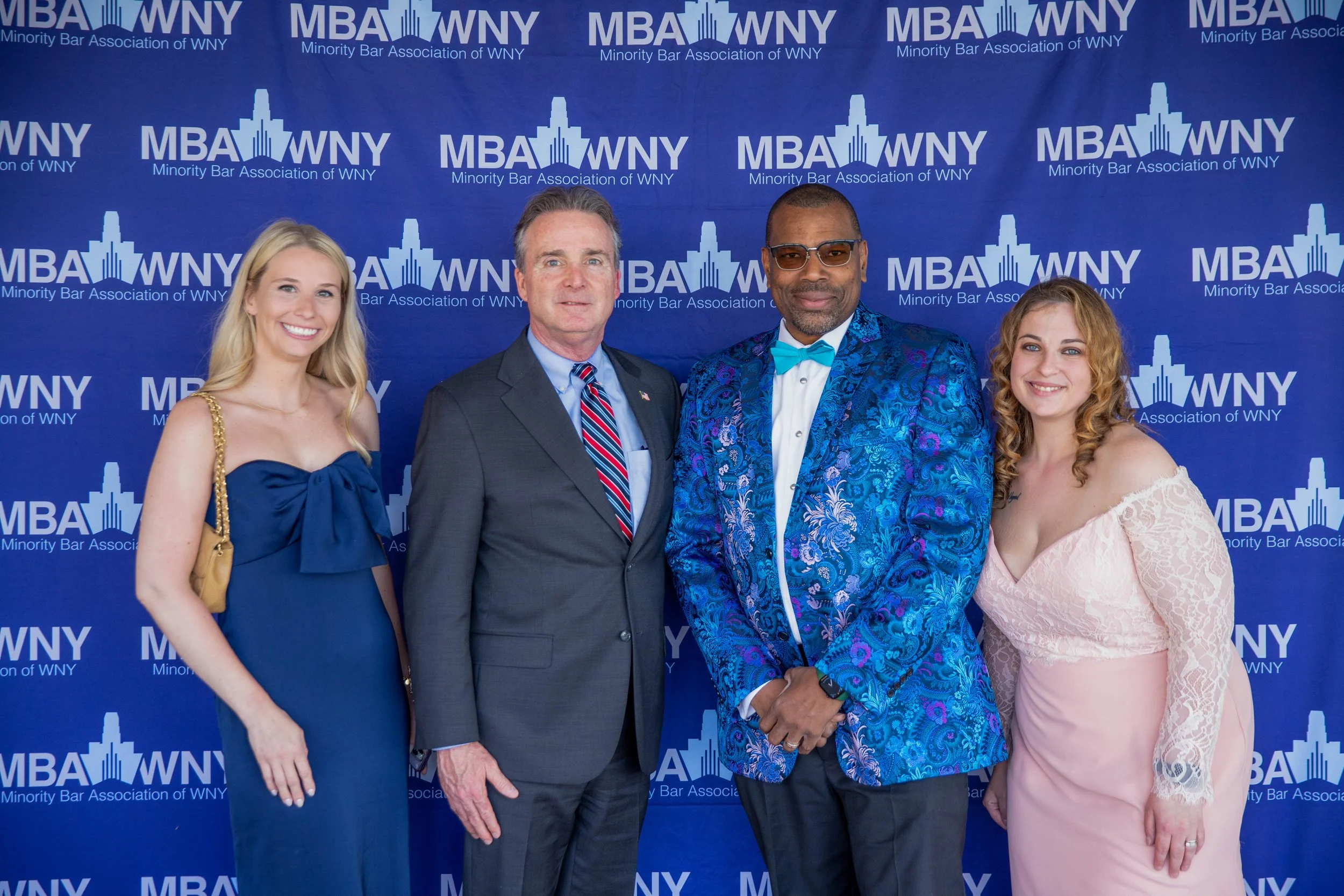 A group of five people standing in front of a blue backdrop with the logos and text of the Minority Bar Association of WNY. The group includes three women and two men, all dressed in formal attire, smiling for the photograph.