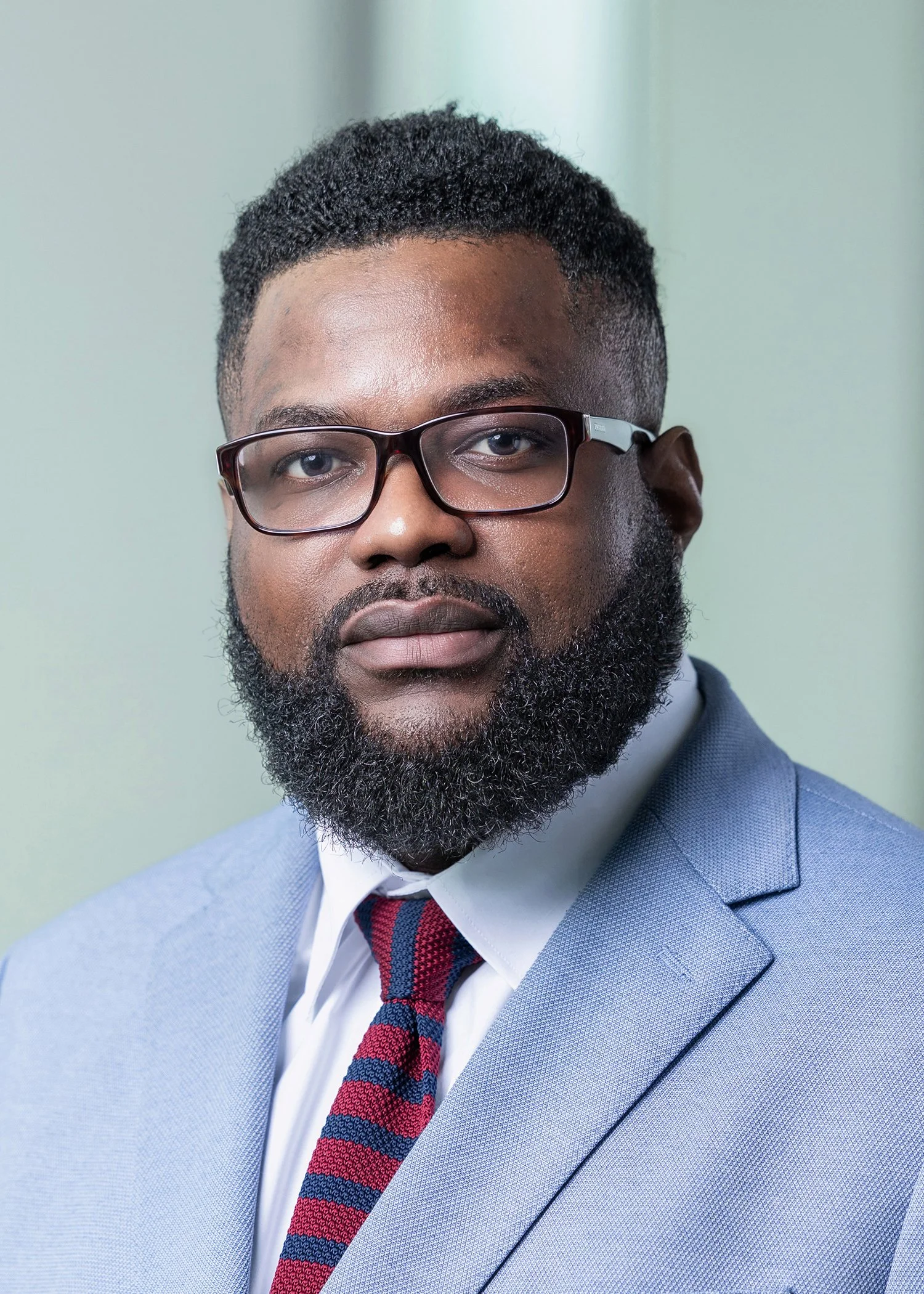 Close-up portrait of a man with dark skin, black glasses, facial hair, dressed in a light blue suit, white shirt, and a red and blue striped tie, against a pale background.