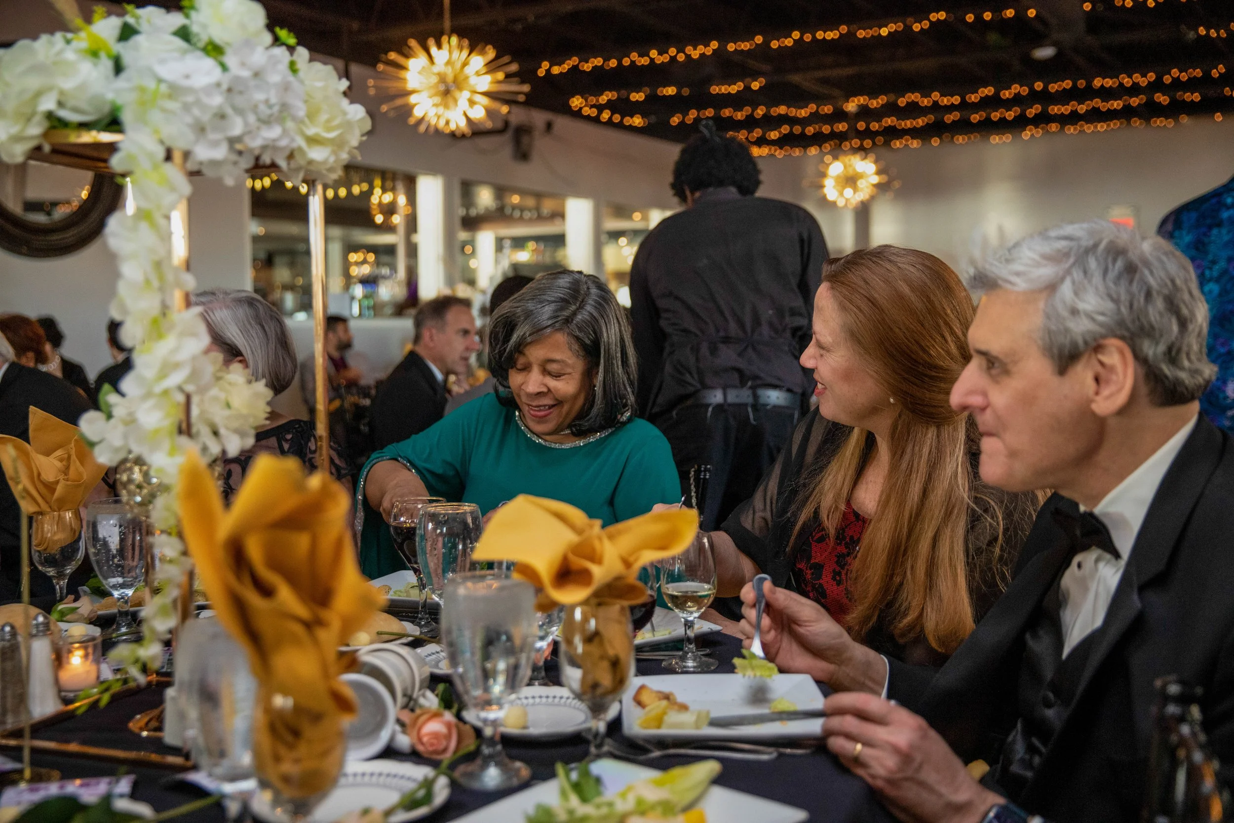 People dressed in formal attire dining at a banquet table decorated with flowers and napkins, with festive lights and hanging spherical light fixtures in the background.