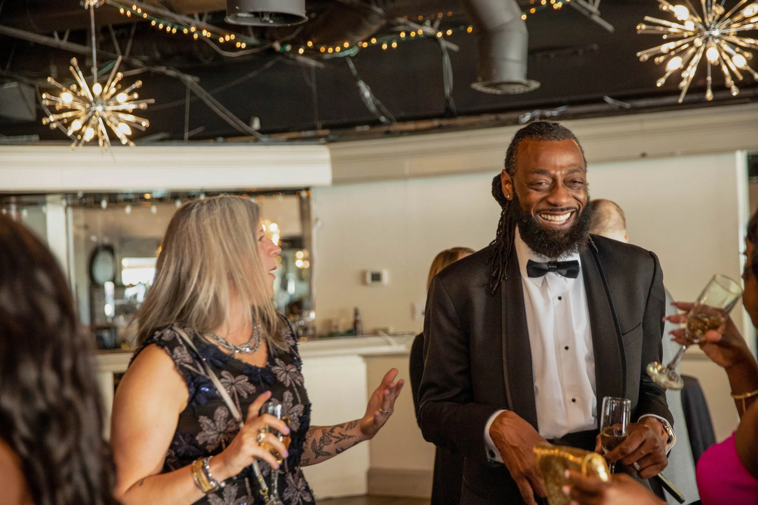 A man in a black tuxedo with a bow tie is smiling and holding a glass, standing at a social gathering with several other people. A woman with tattoos and jewelry is talking to him, while other guests hold drinks and socialize. The setting appears to 