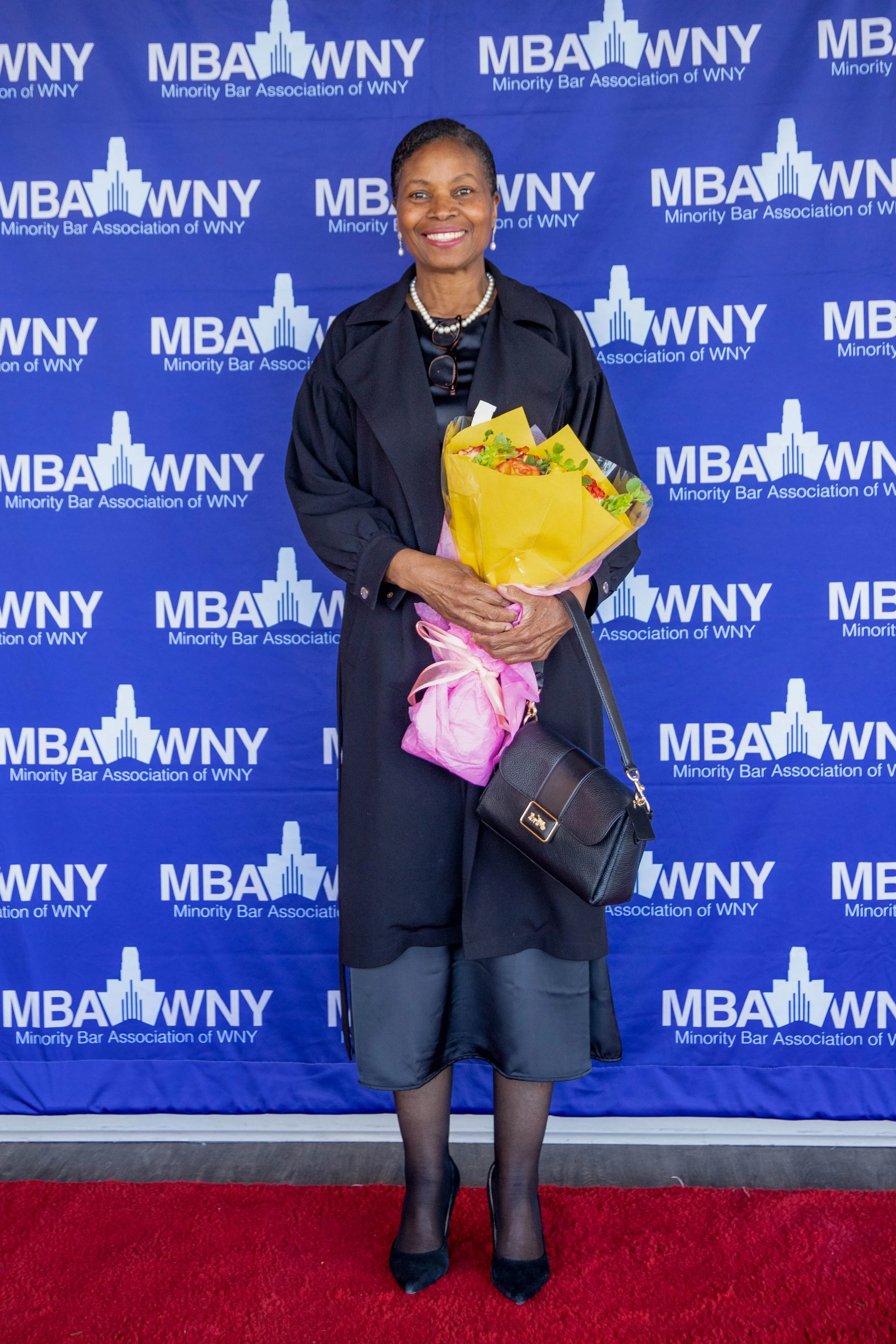 A woman in a black judge's robe holding a bouquet of flowers on a red carpet, standing in front of a blue backdrop with the logo and text reading "Minority Bar Association of WNY".