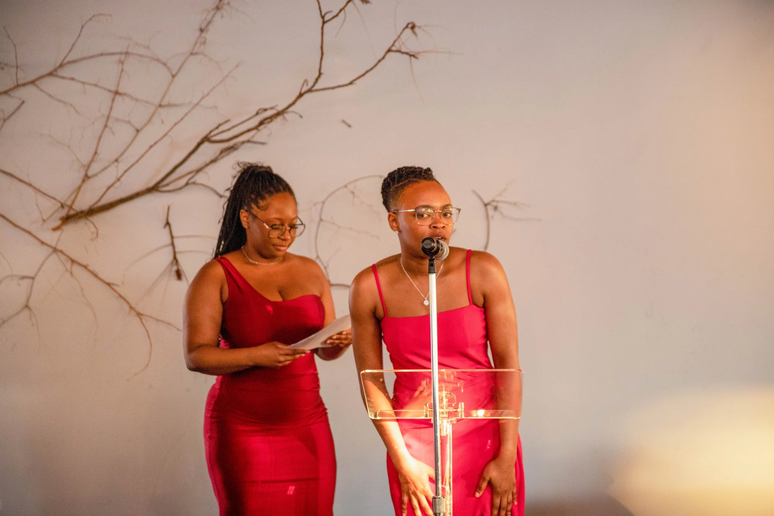 Two women in red dresses standing behind a clear podium, one speaking into a microphone, with a wall decorated with bare tree branches in the background.