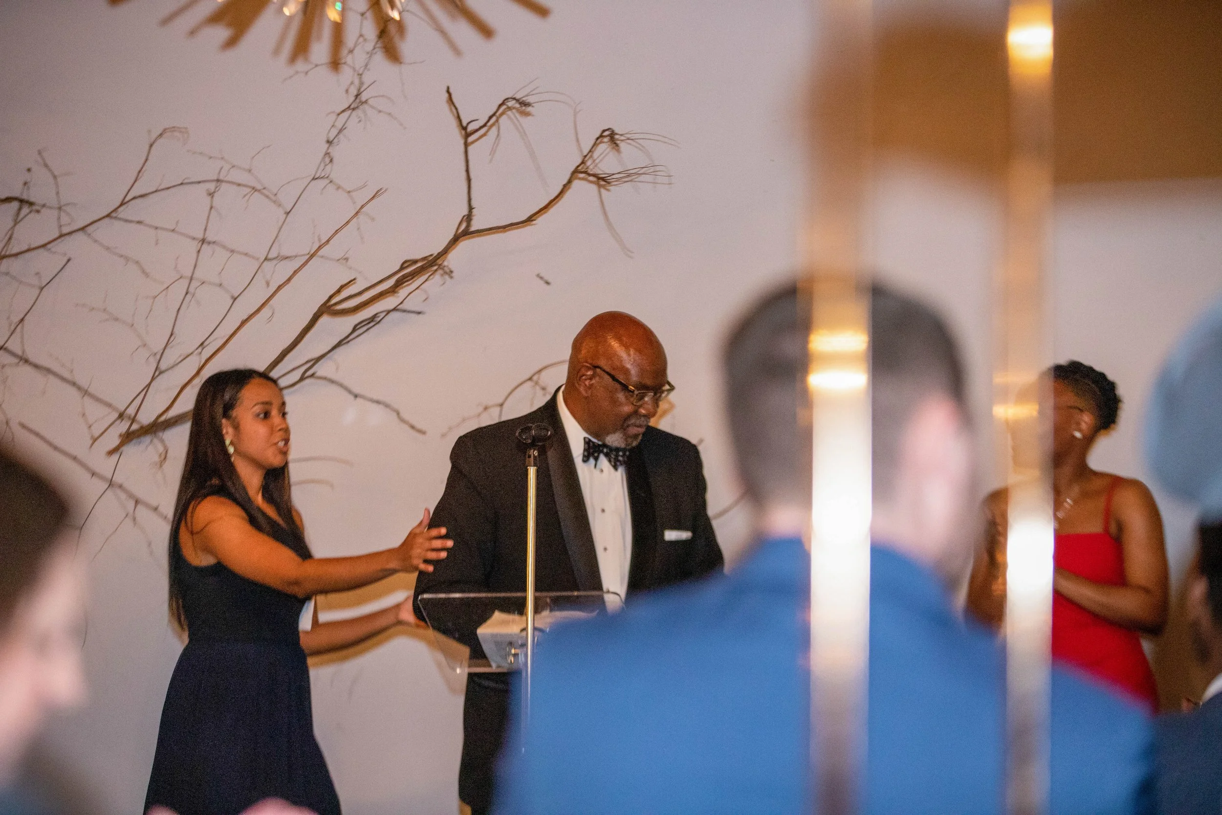 A man in a tuxedo and glasses stands at a podium with a microphone, while a woman in a black dress touches his arm. Other women in red dresses are visible in the background at a formal event.