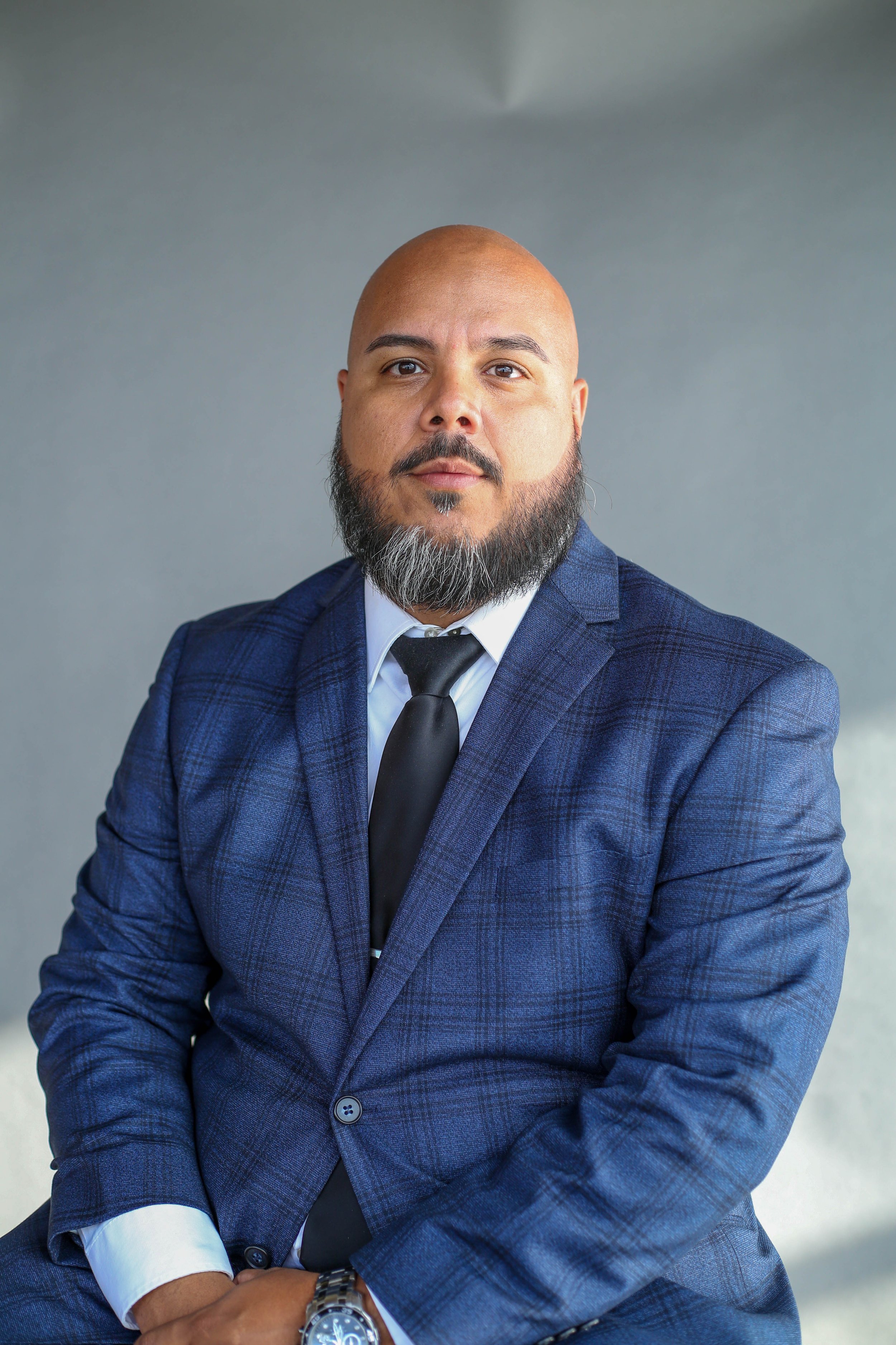 A man with a bald head and a beard wearing a blue plaid suit, white shirt, and black tie, sitting with arms crossed against a gray background.