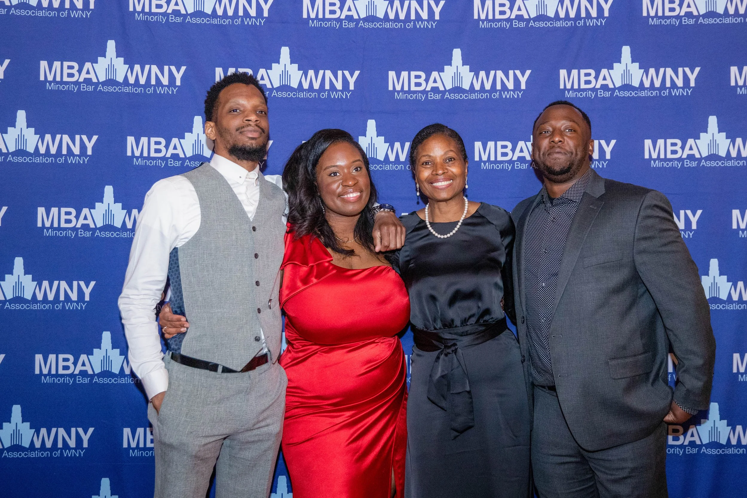 Four individuals dressed in formal attire standing together in front of a blue backdrop with the logo and text of the Minority Bar Association of WNY, posing for a group photo at an event.